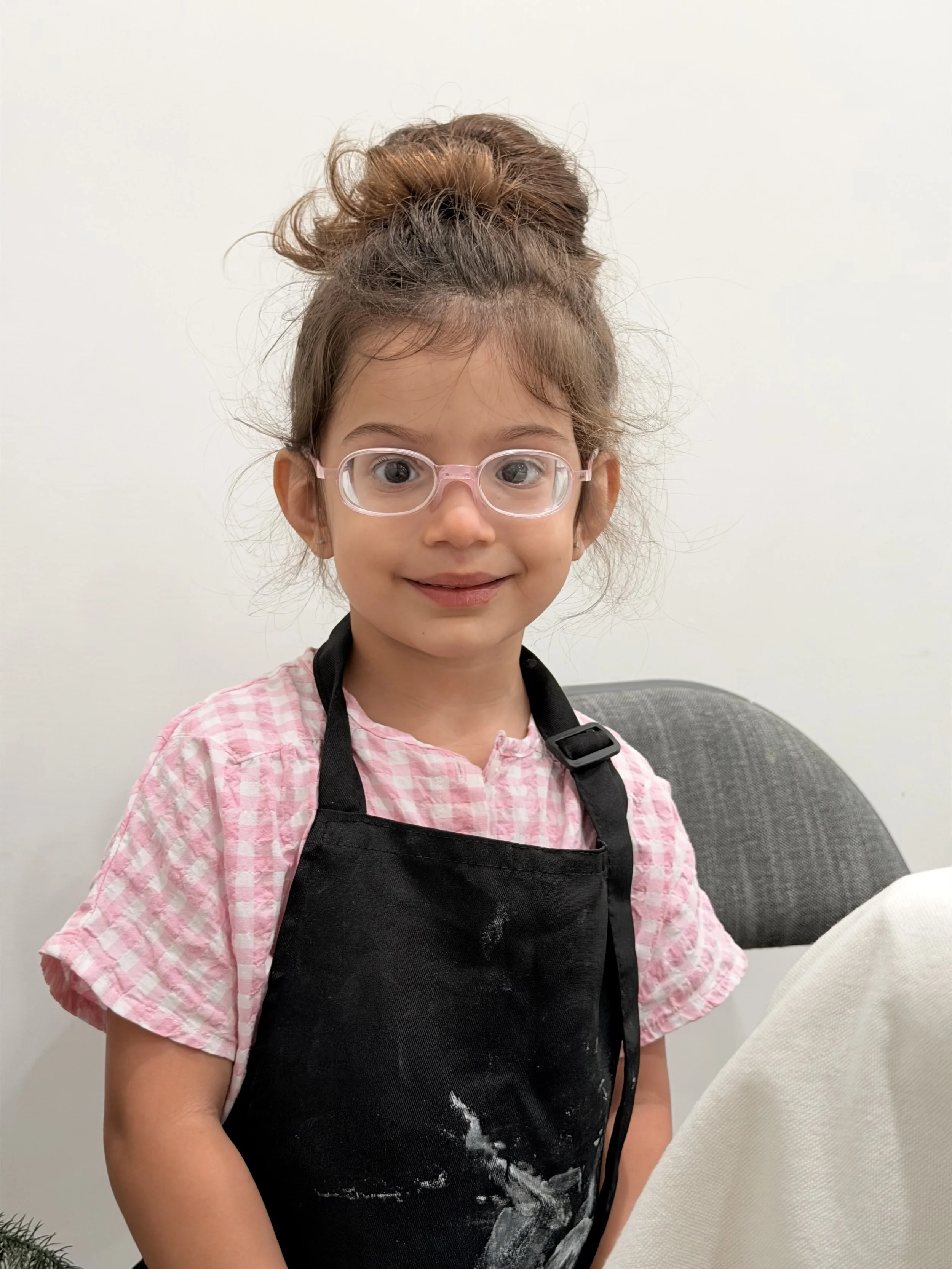 A young girl with glasses, wearing a pink checkered shirt and a black apron, standing against a plain white wall.