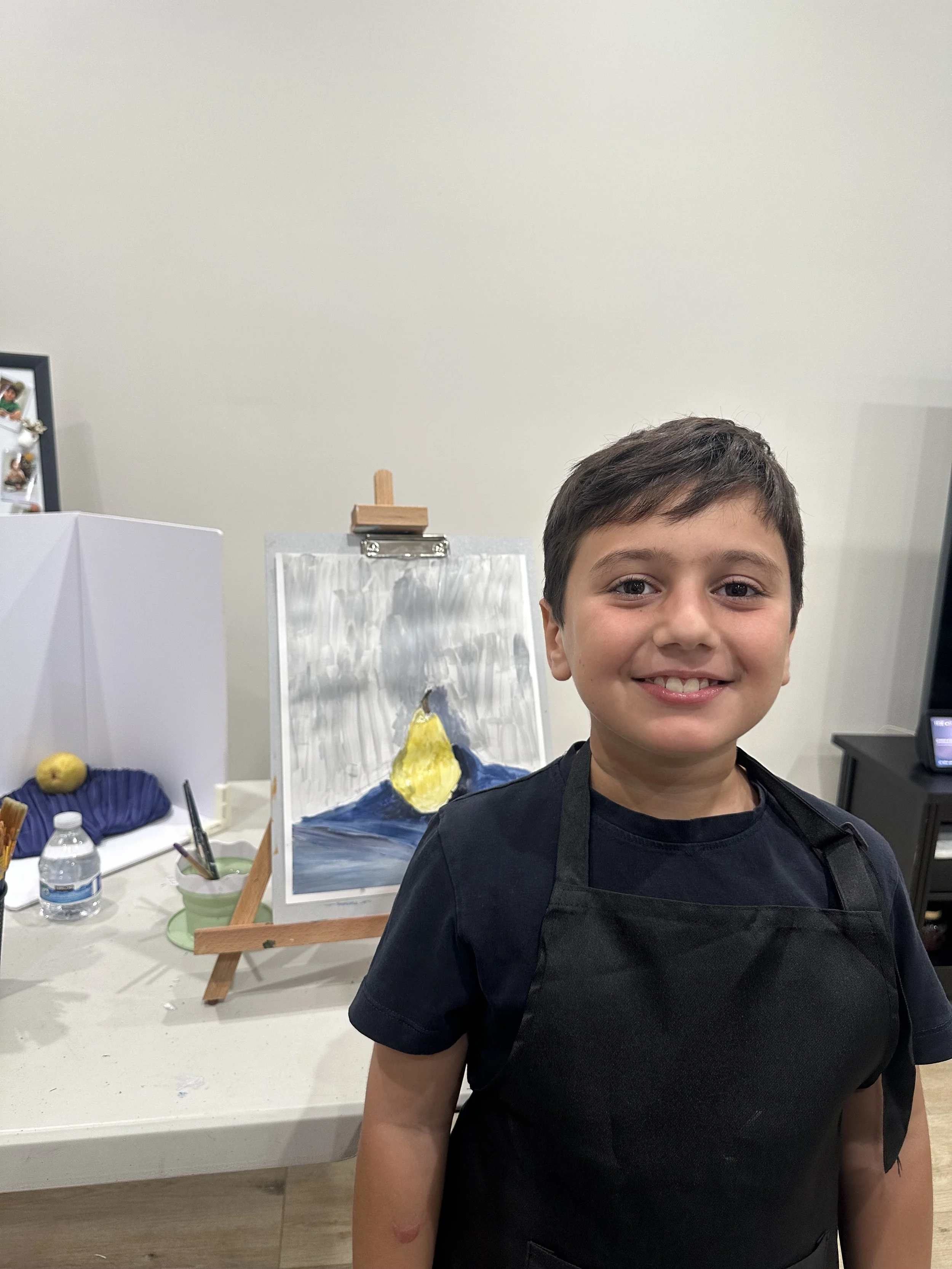 A young boy smiling, wearing a black apron and standing in front of a table with art supplies and a painting of a mountain landscape with a yellow sun.