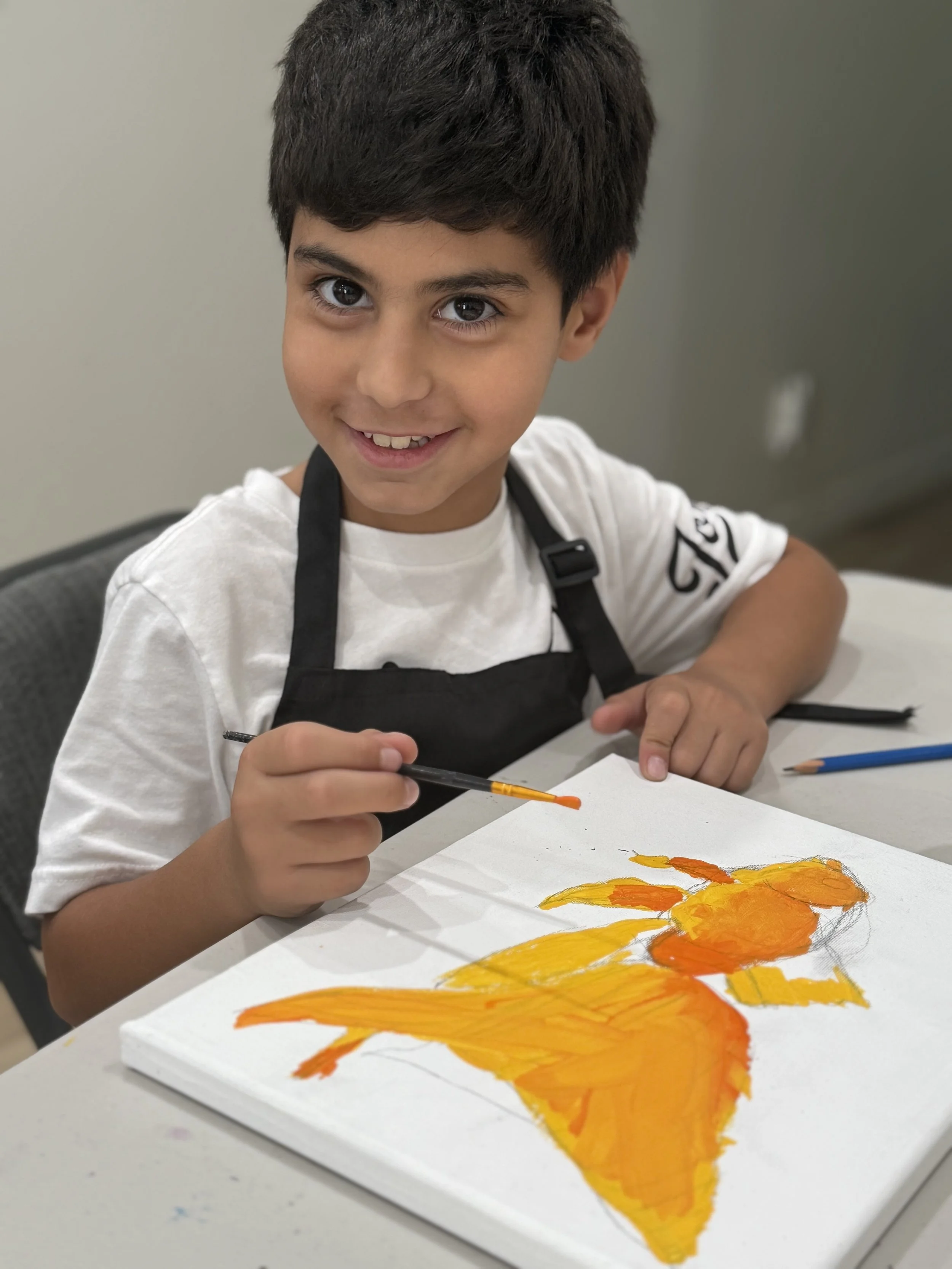 A young boy with dark hair, wearing a white shirt and black apron, is sitting at a table painting a picture with orange and yellow colors on a white canvas. He is smiling and looking at the camera.