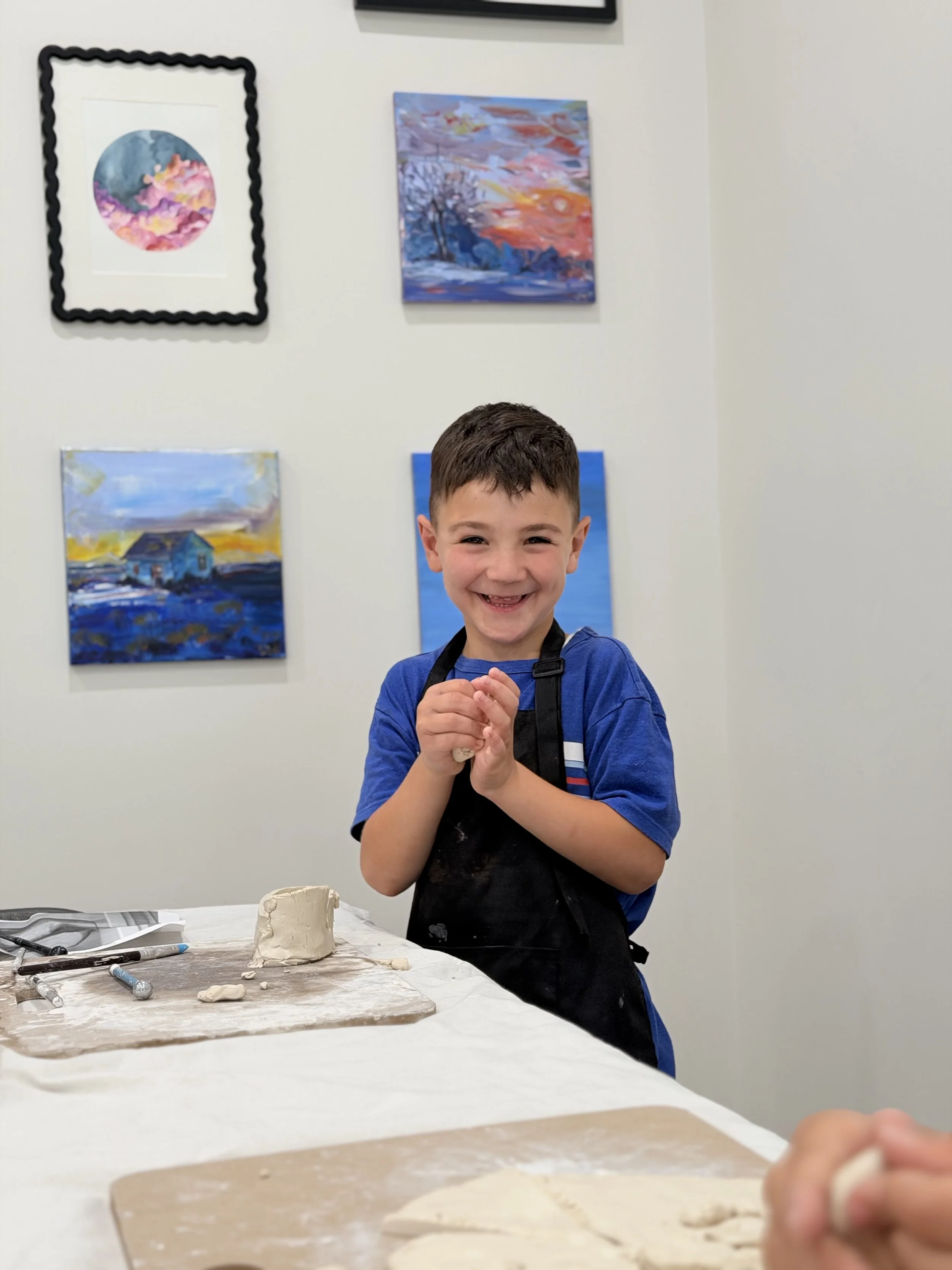 A smiling young boy in a blue shirt and black apron stands in front of a table with clay and sculpting tools, with colorful paintings on the wall behind him.