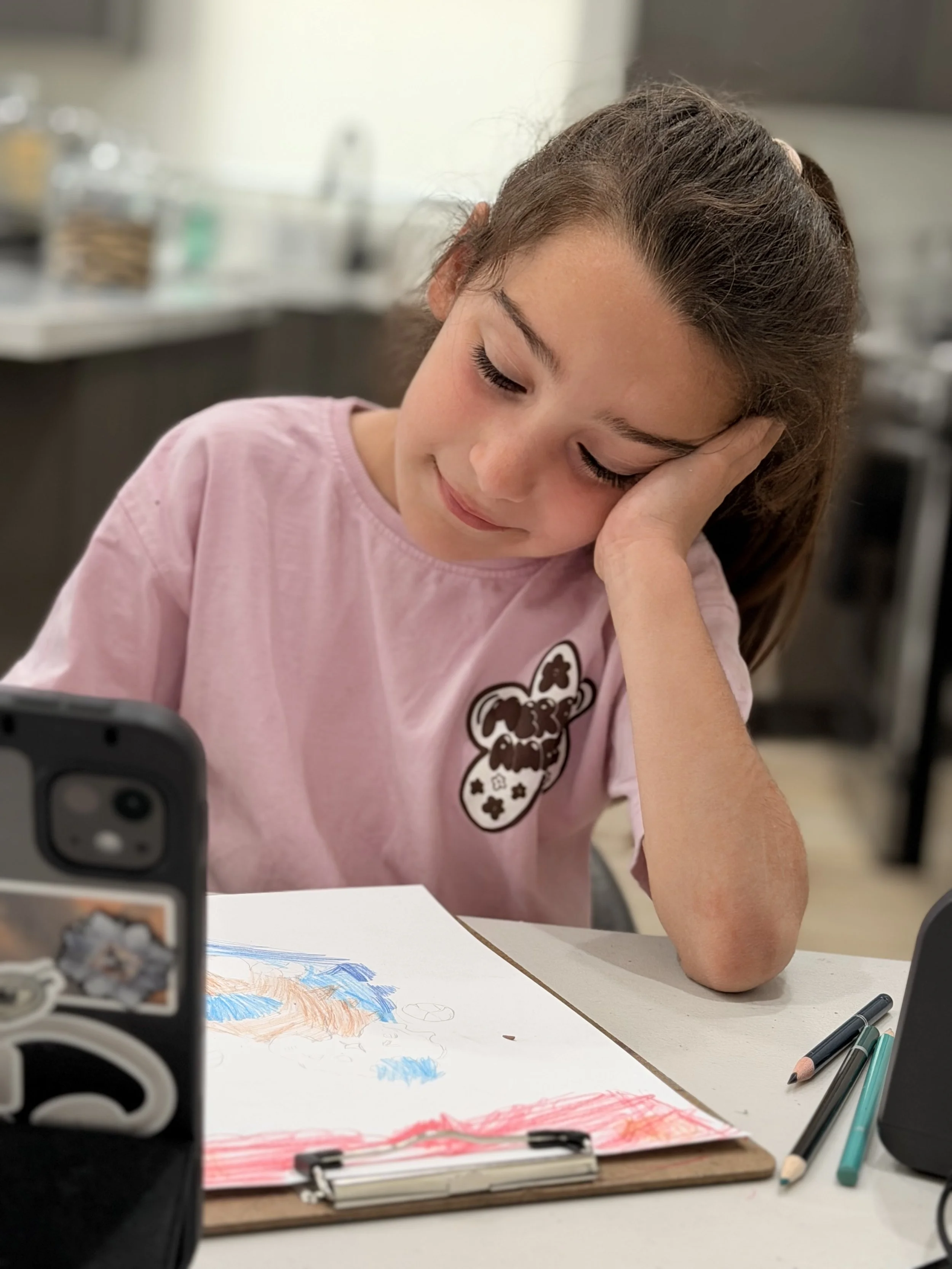 A young girl with long brown hair tied in a ponytail, wearing a pink shirt, is resting her head on her hand while looking down. She is sitting at a table with colored markers and a drawing or coloring project in front of her. The background is a blurred kitchen or workspace.