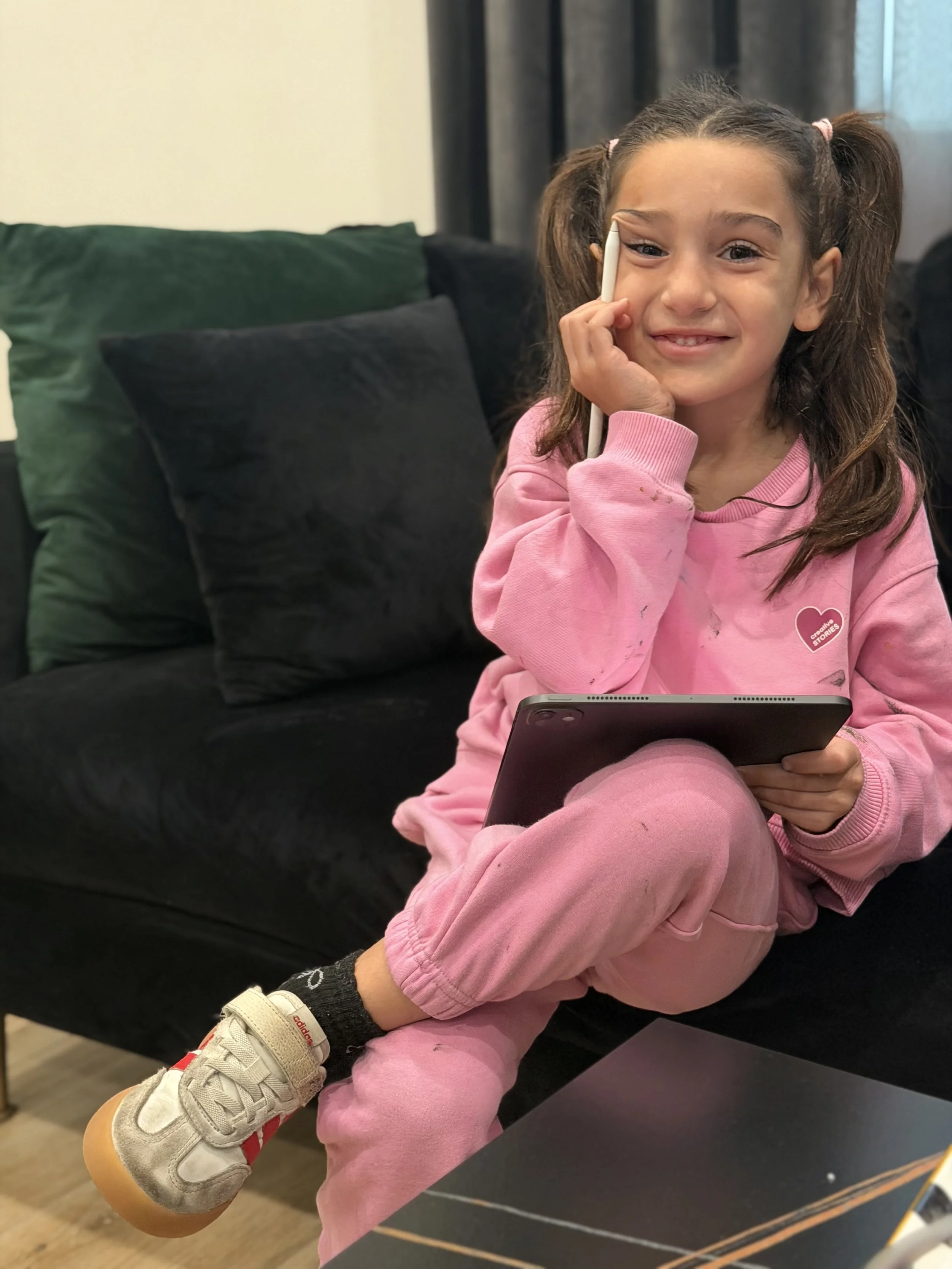A young girl with brown hair in pigtails, wearing a pink sweatshirt and matching pants, sitting on a black couch, holding a tablet in her lap, smiling, with her right hand touching her face and a stylus in her left hand.