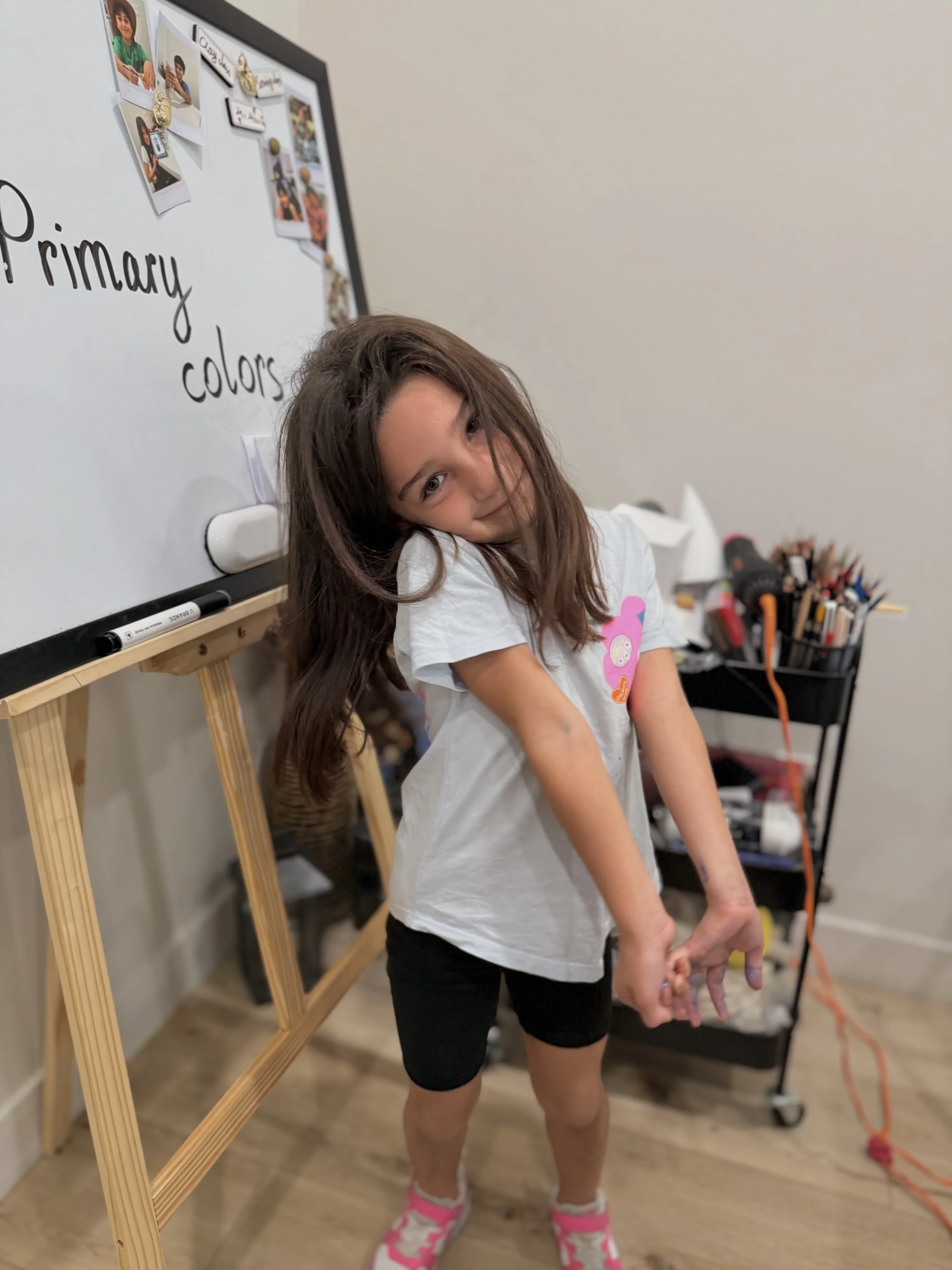 A young girl with long brown hair and a white t-shirt standing in front of a whiteboard with the words 'Primary colors' written on it. She has a slight smile and her head tilted to the side, with her arms extended downward. There is a container with 