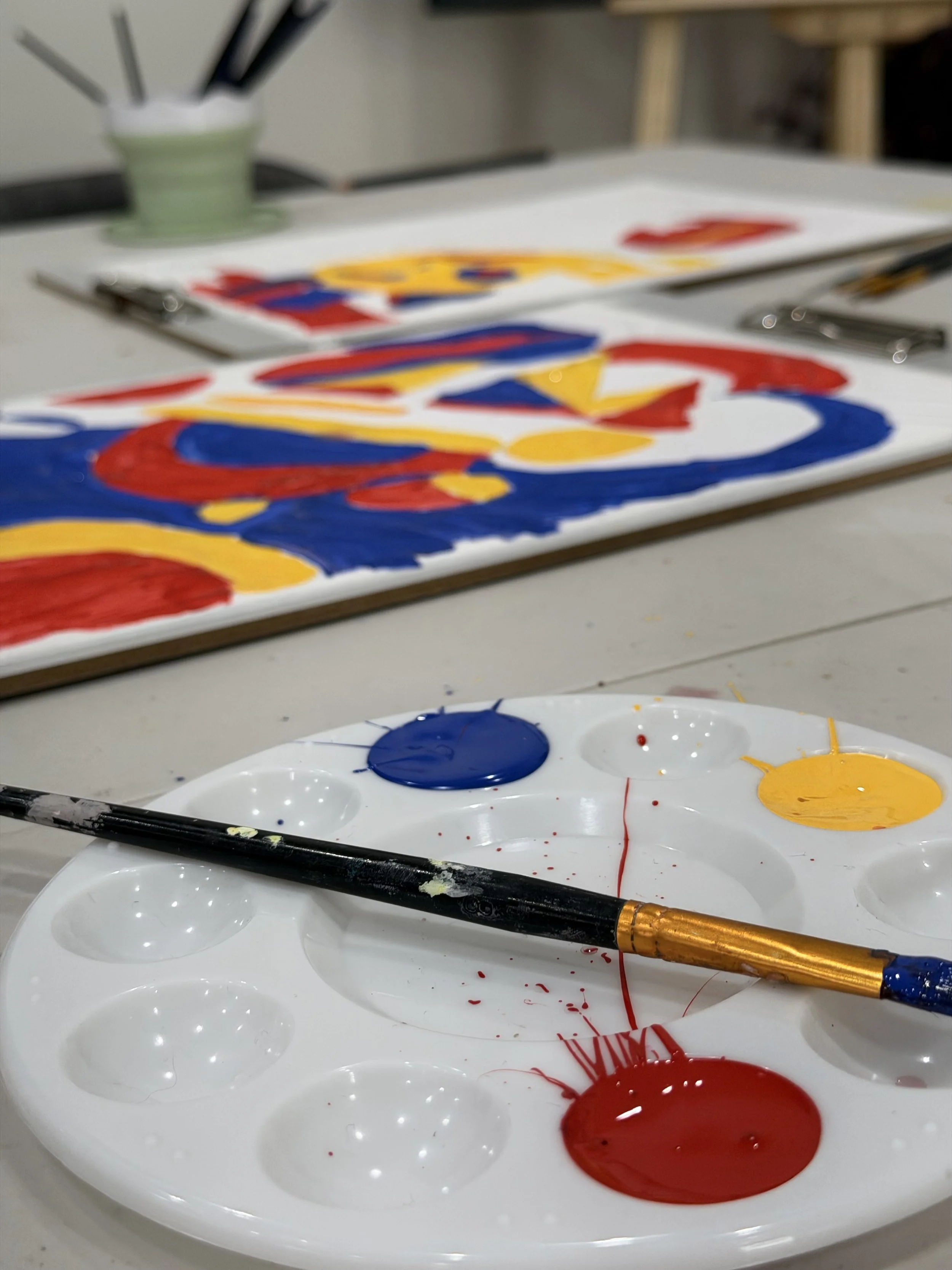Paint palette with blue, yellow, and red paint, and a brush with blue paint resting on it, in an art studio.