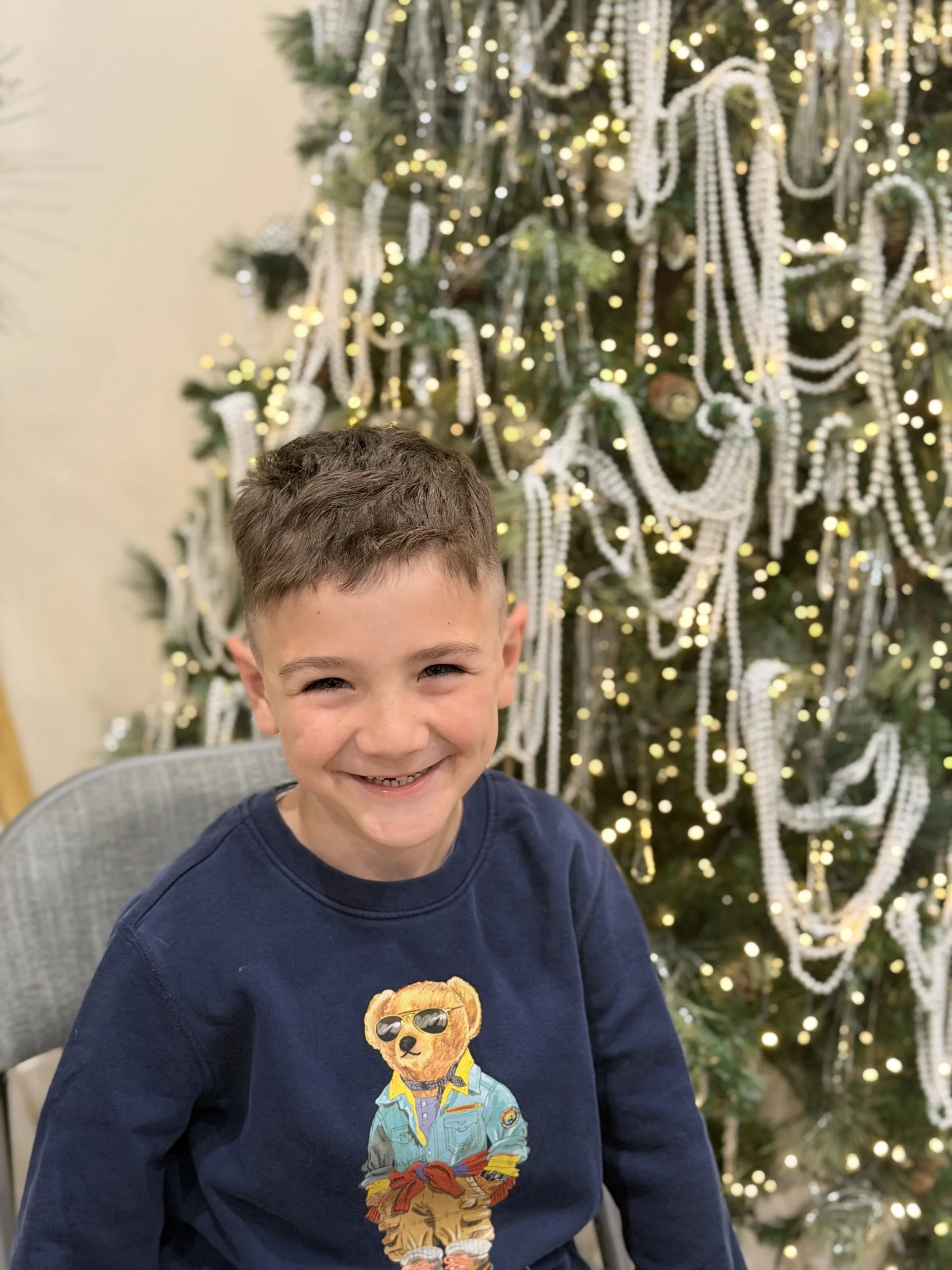 A young boy with short brown hair and a big smile, wearing a navy blue sweat shirt with a graphic of a bear dressed as a cowboy, sitting in front of a decorated Christmas tree with lights and white bead garlands.