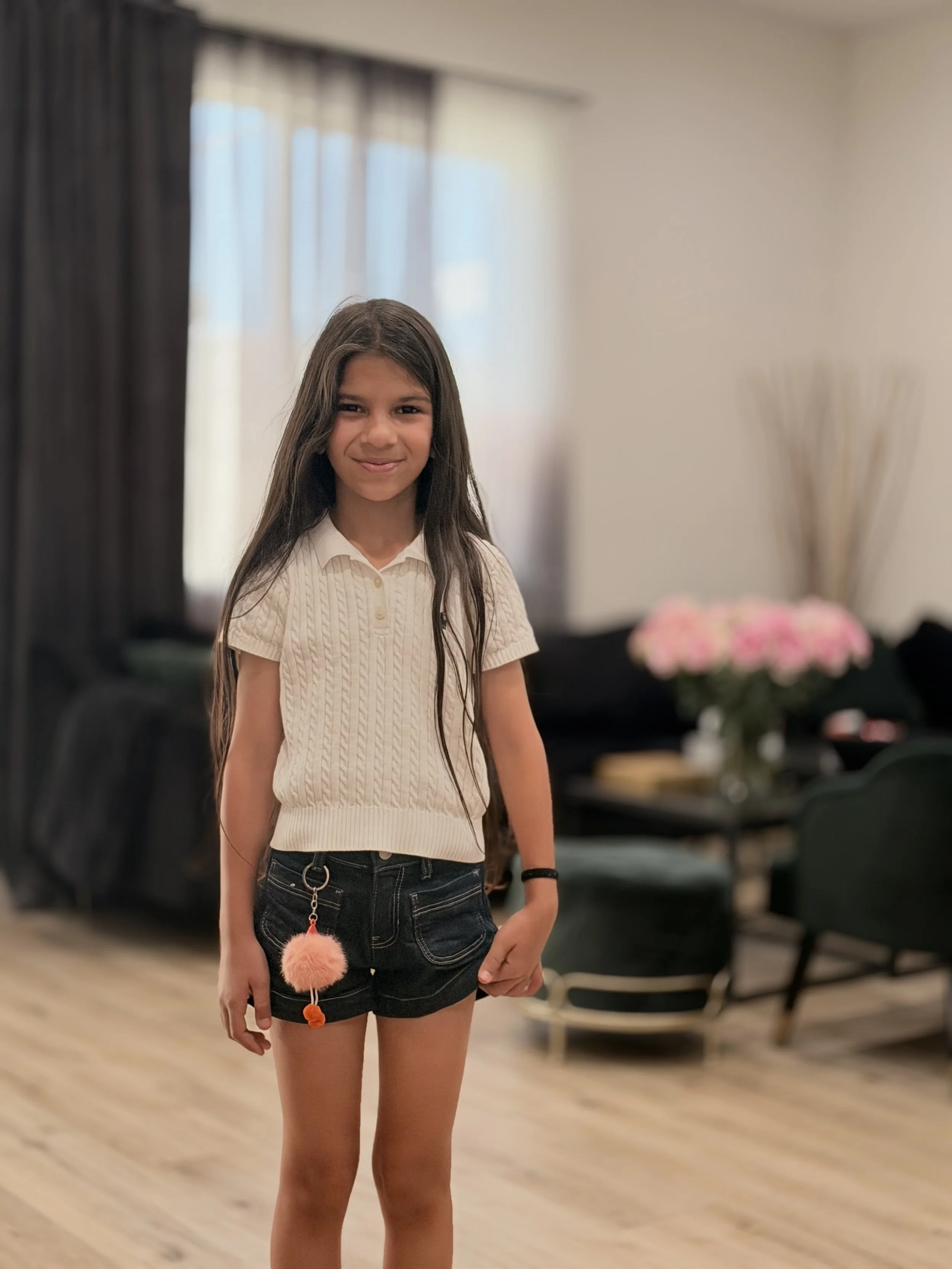 A young girl with long dark hair, wearing a cream-colored short-sleeve sweater, black shorts, and a black wristband, stands inside a room with a wooden floor, blurred background of tables and chairs, a window with curtains, and pink flowers on a tabl