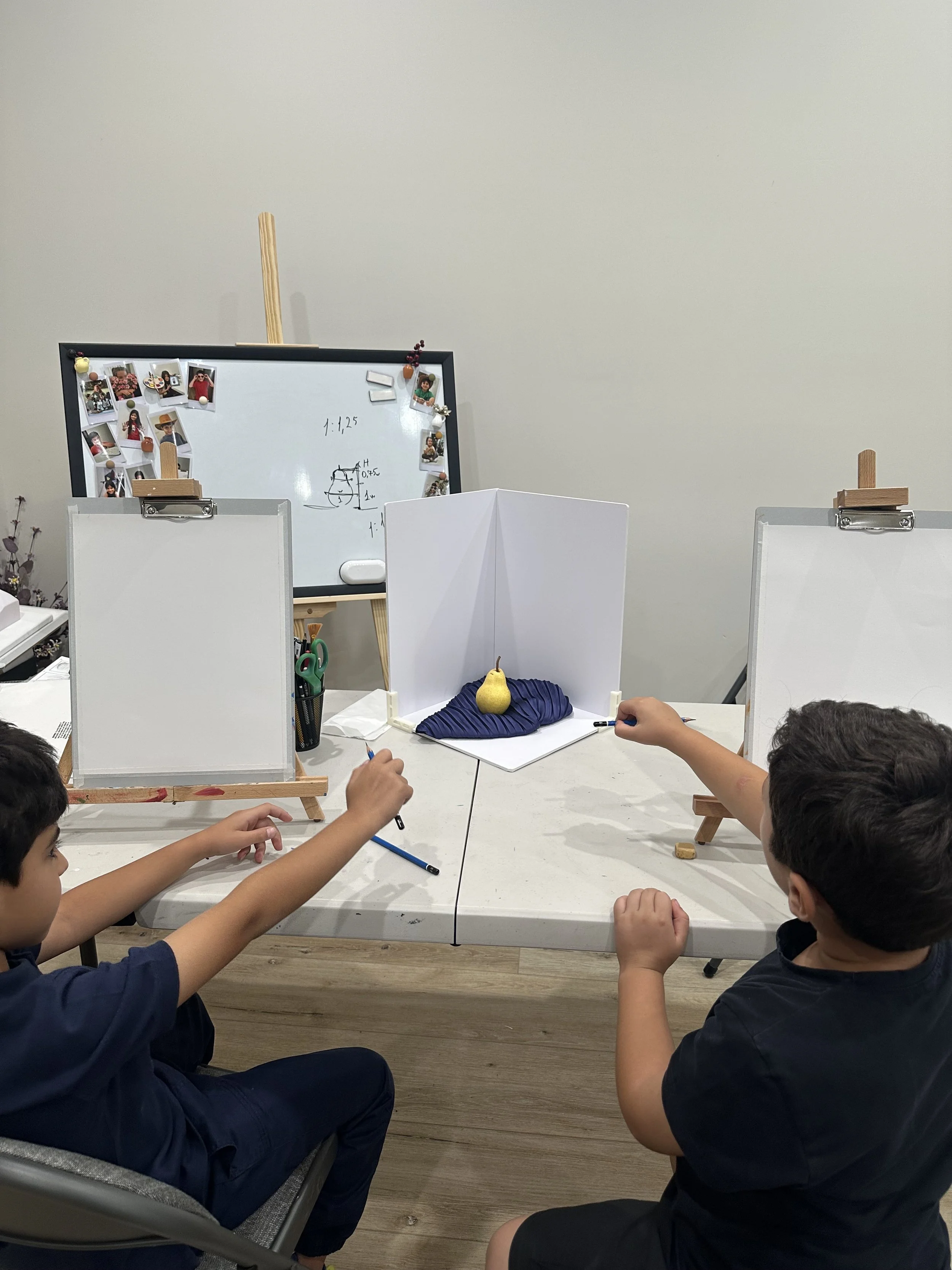 Three children sitting at a table playing a game involving a pear and a blue fabric, with white dividers and a whiteboard with photos and notes in the background.