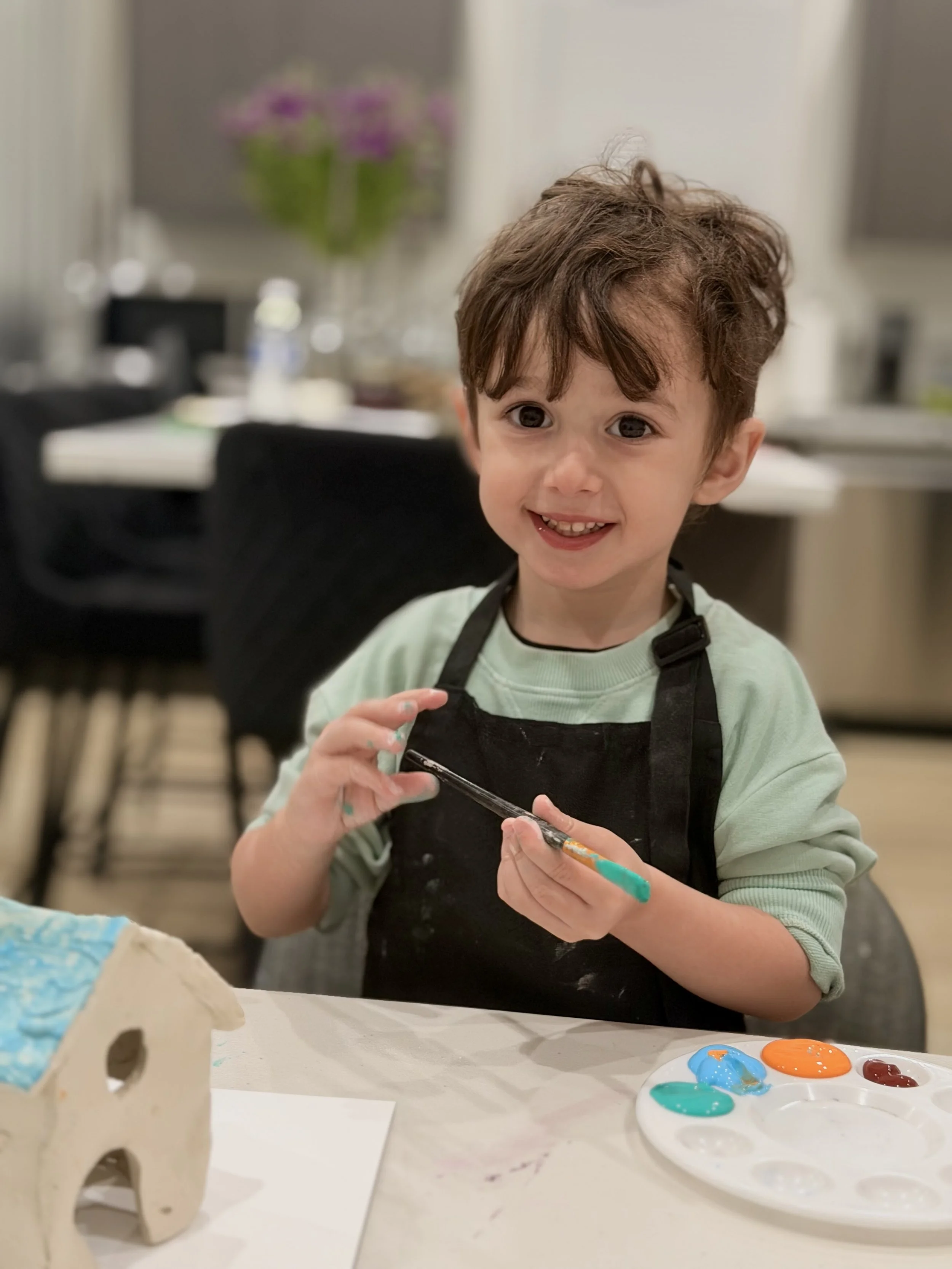 A young boy with curly brown hair and big eyes smiling while holding a paintbrush. He is wearing a black apron over a light green shirt. In front of him on the table, there's a palette with various bright paint colors, and a partially painted ceramic