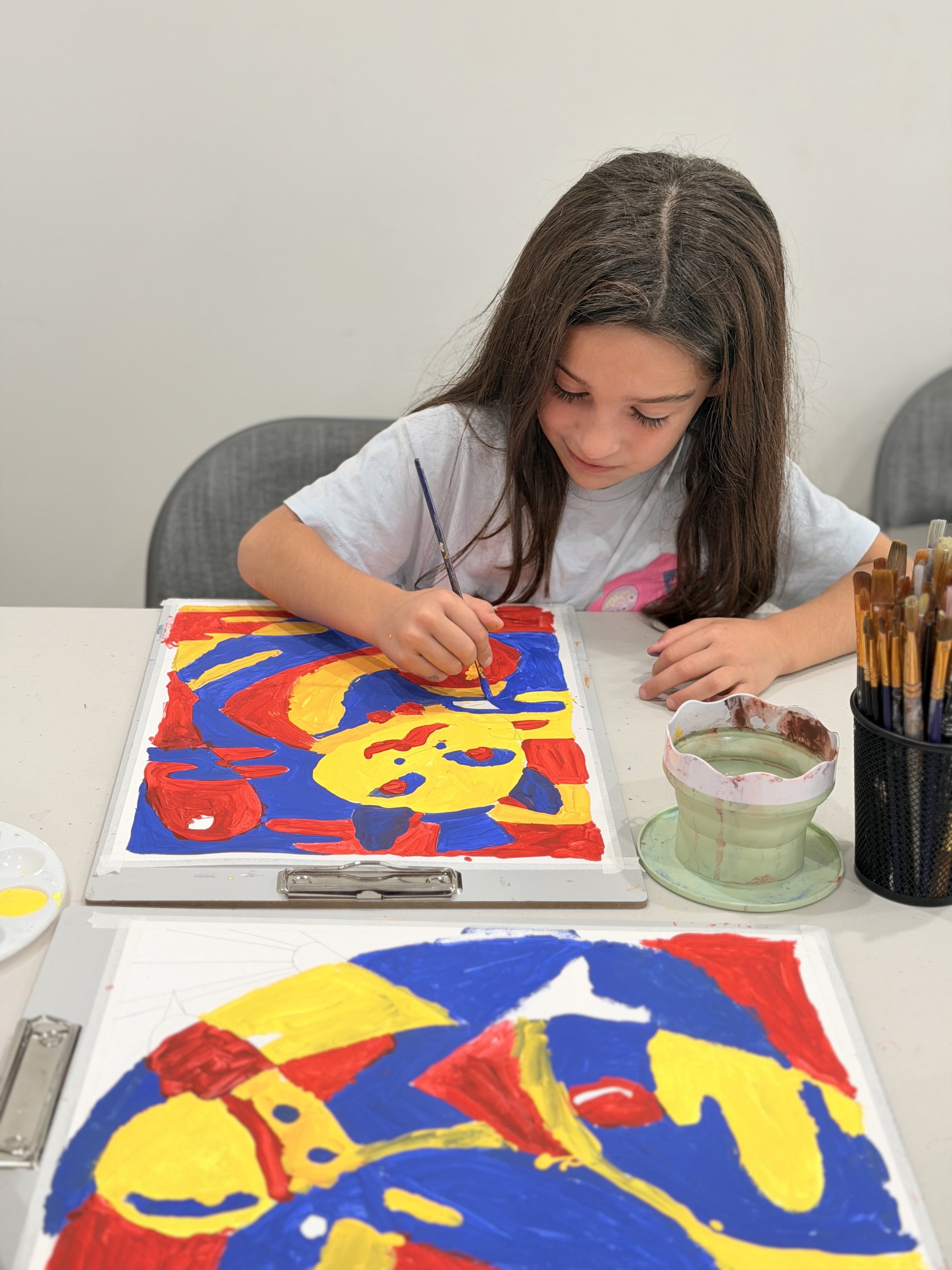 A young girl with long brown hair painting an abstract artwork with red, blue, and yellow colors on a white paper, seated at a white table with art supplies around her.