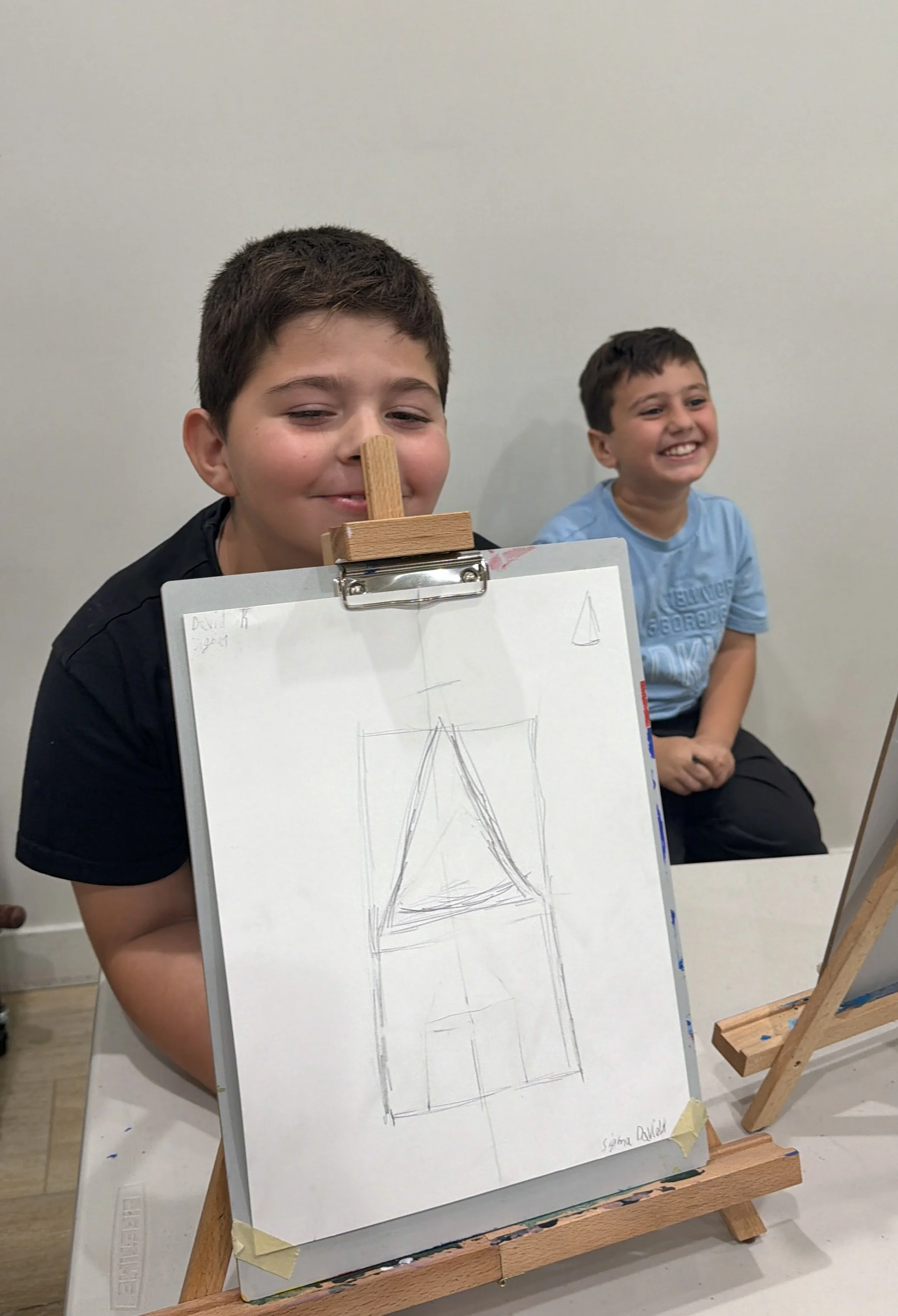 Two young boys sitting in front of an easel with a paper sketch of the Eiffel Tower. One boy is close to the camera, smiling and wearing a black shirt, while the other boy is in the background, smiling and wearing a blue shirt.