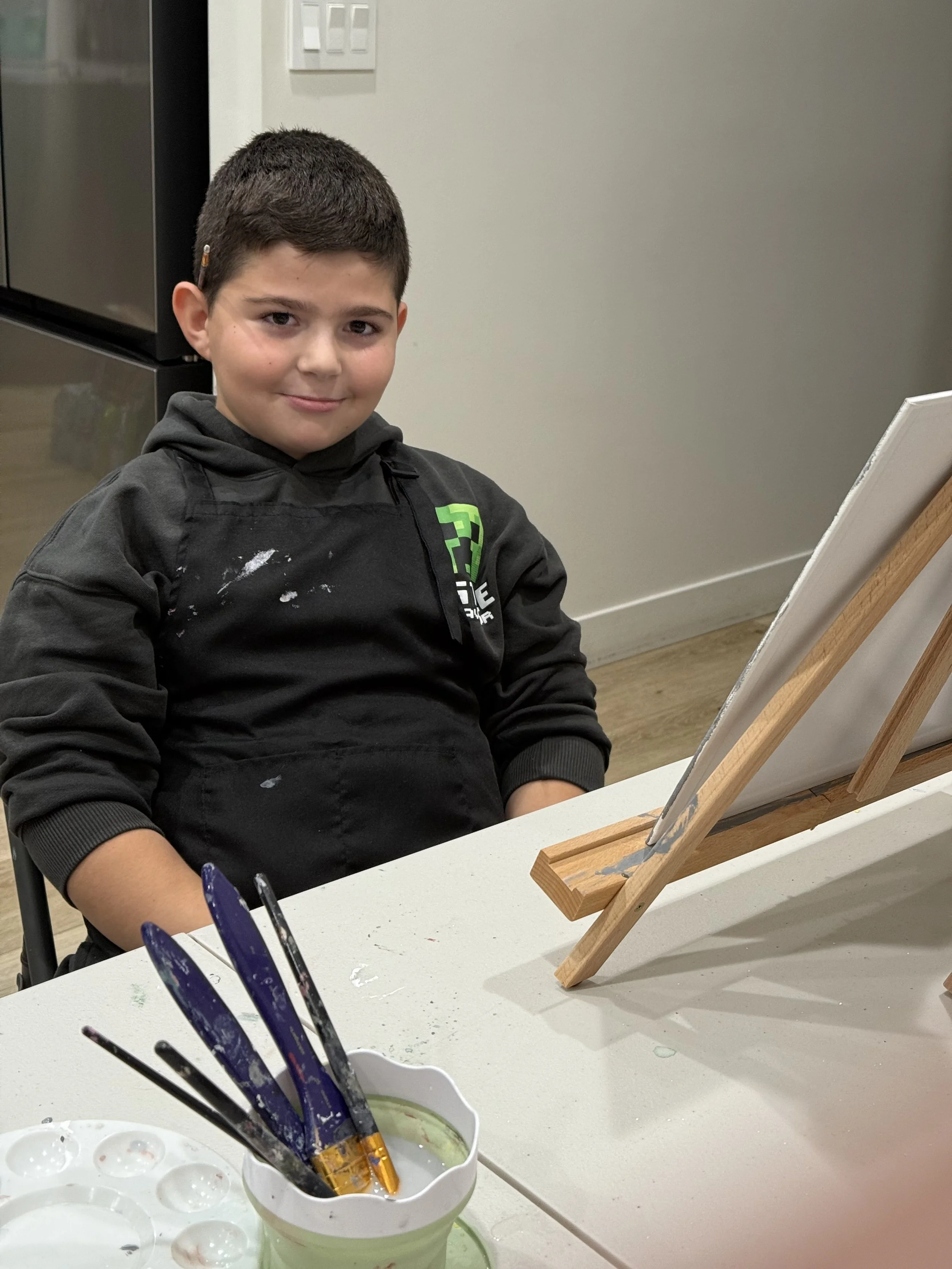 A young boy sitting at a table with paint brushes in a cup, and a small wooden easel with blank canvas in front of him, in a room with a gray wall and light switches.