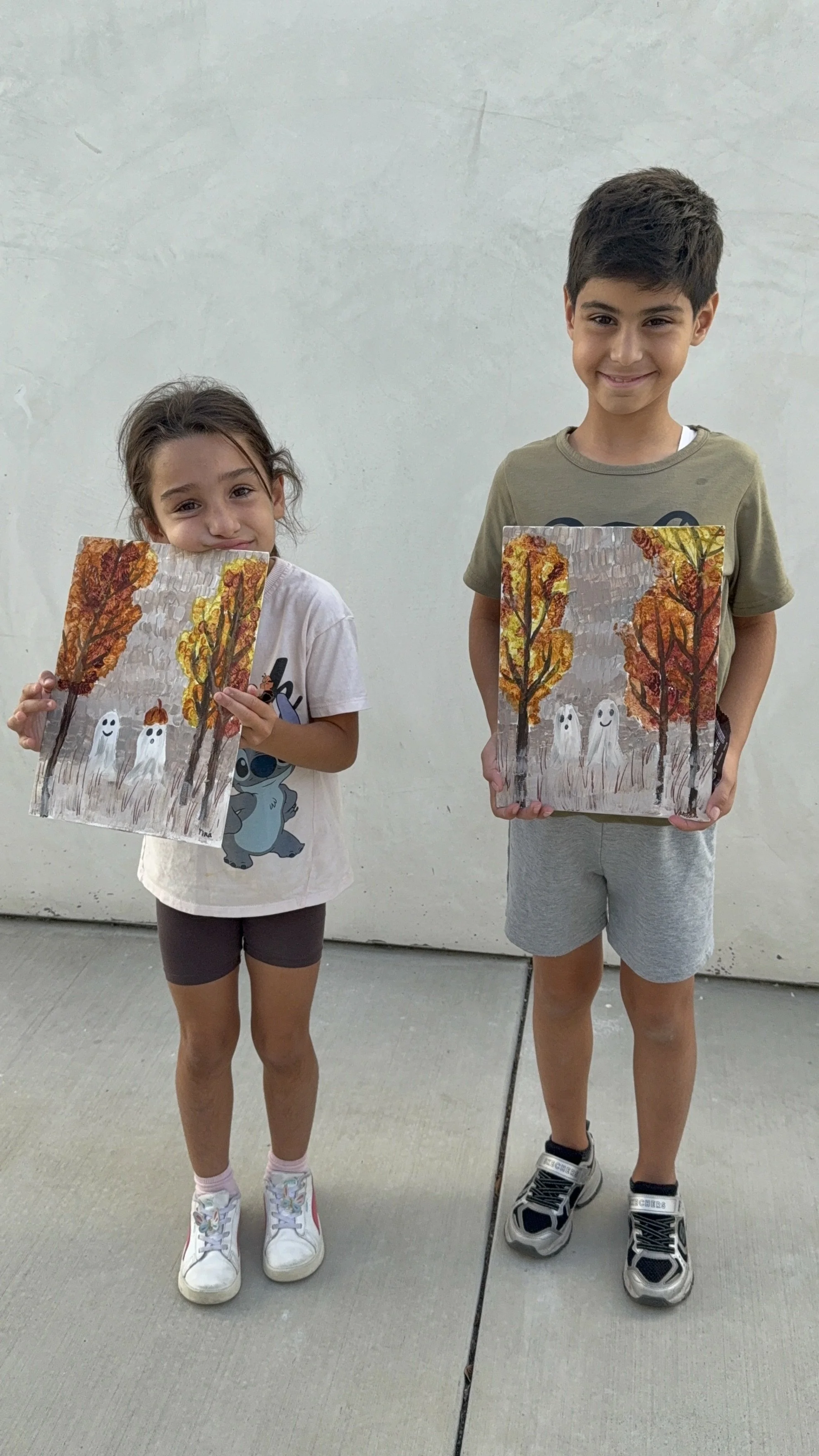 Young girl and boy standing on a sidewalk, each holding a canvas with fall-themed artwork of trees and white ghost-like figures.