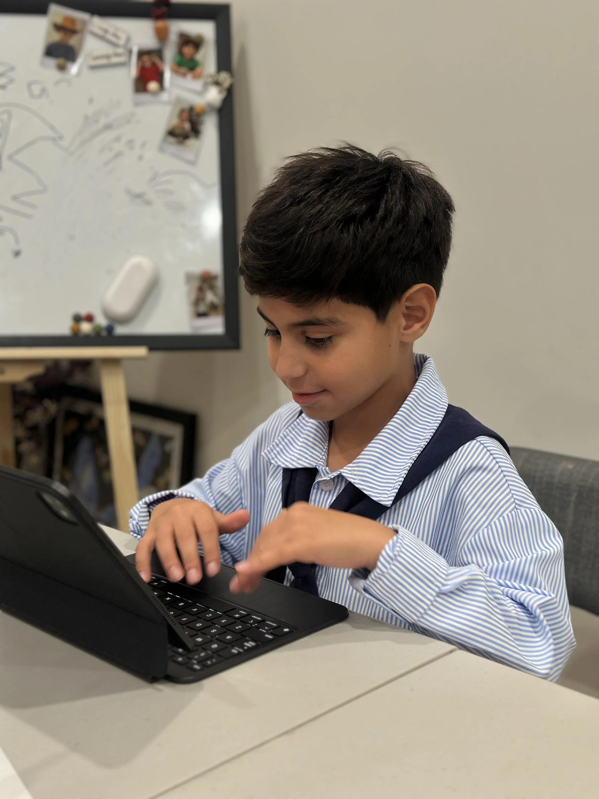 A young boy with short dark hair wearing a blue and white striped shirt, sitting at a table, typing on a black keyboard connected to a computer. In the background, there is a whiteboard with photos and drawings, and a small wooden table with some photo frames underneath.