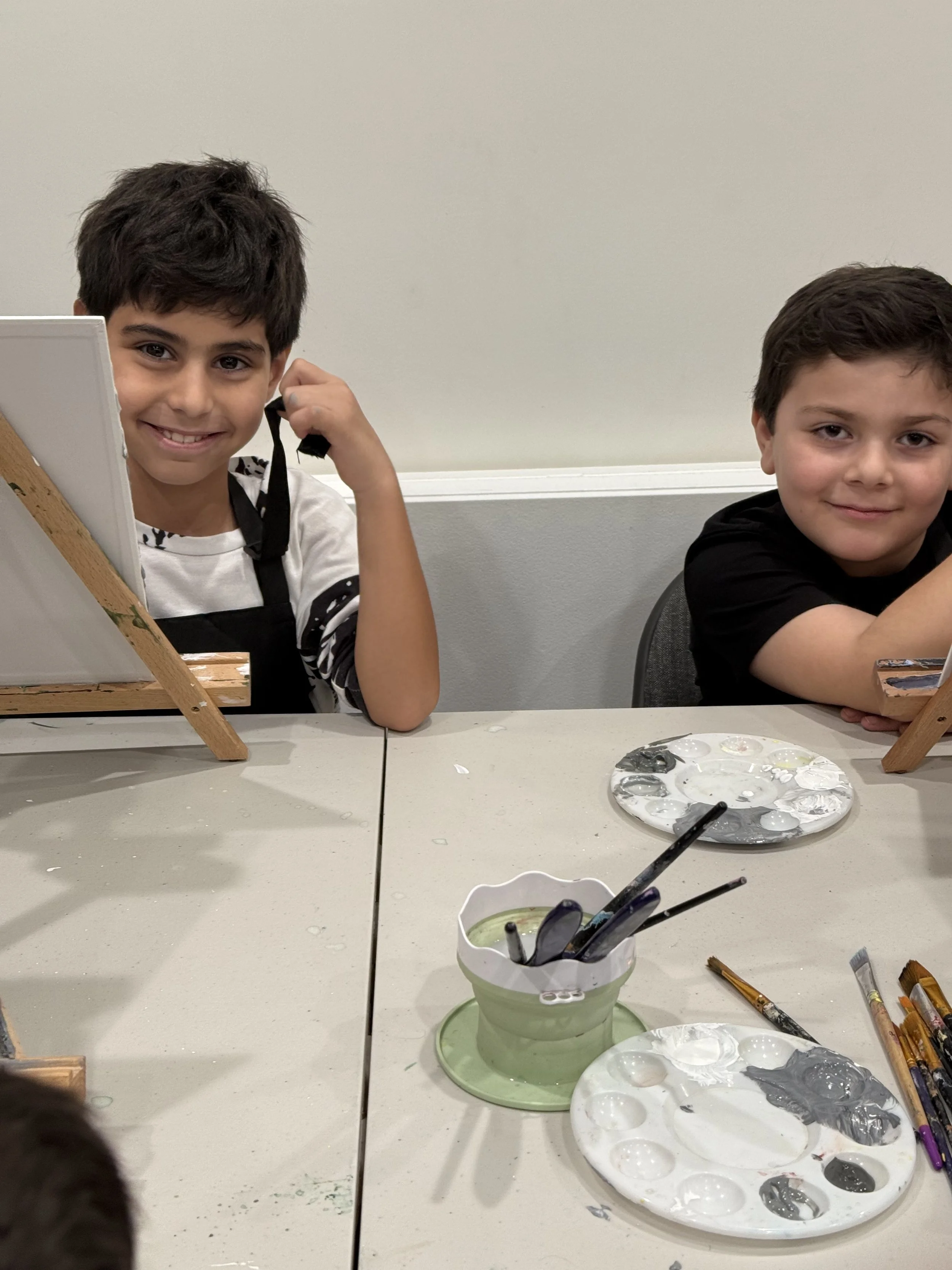 Two young boys sit at a table with art supplies; one is smiling and adjusting his shirt, and the other is resting his arms on the table, both looking at the camera. Art palettes with paint and brushes are on the table in front of them.