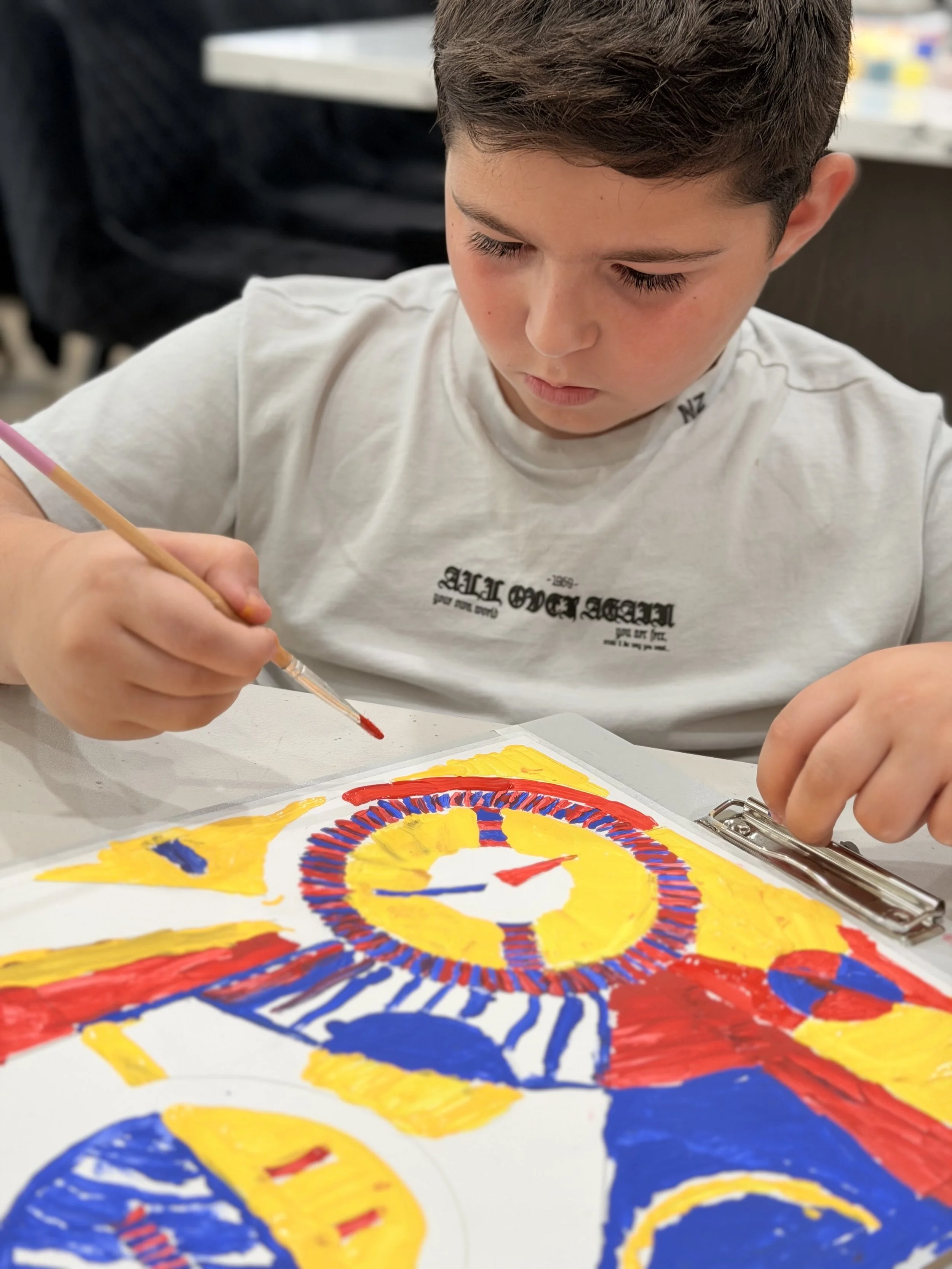 A young boy with short brown hair is painting a colorful abstract design on a white paper with a small paintbrush. The design includes yellow, red, and blue colors, arranged in circular patterns. The boy is focused on his artwork, sitting at a table 