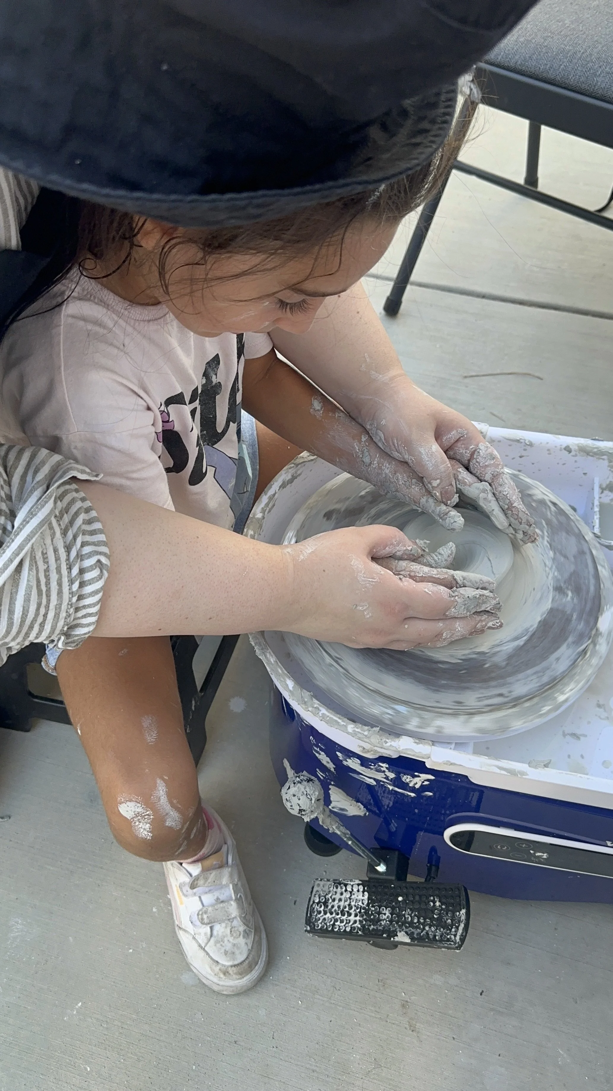 Child shaping clay on a pottery wheel outdoor, hands covered in white clay, wearing sneakers with some clay smeared on legs.