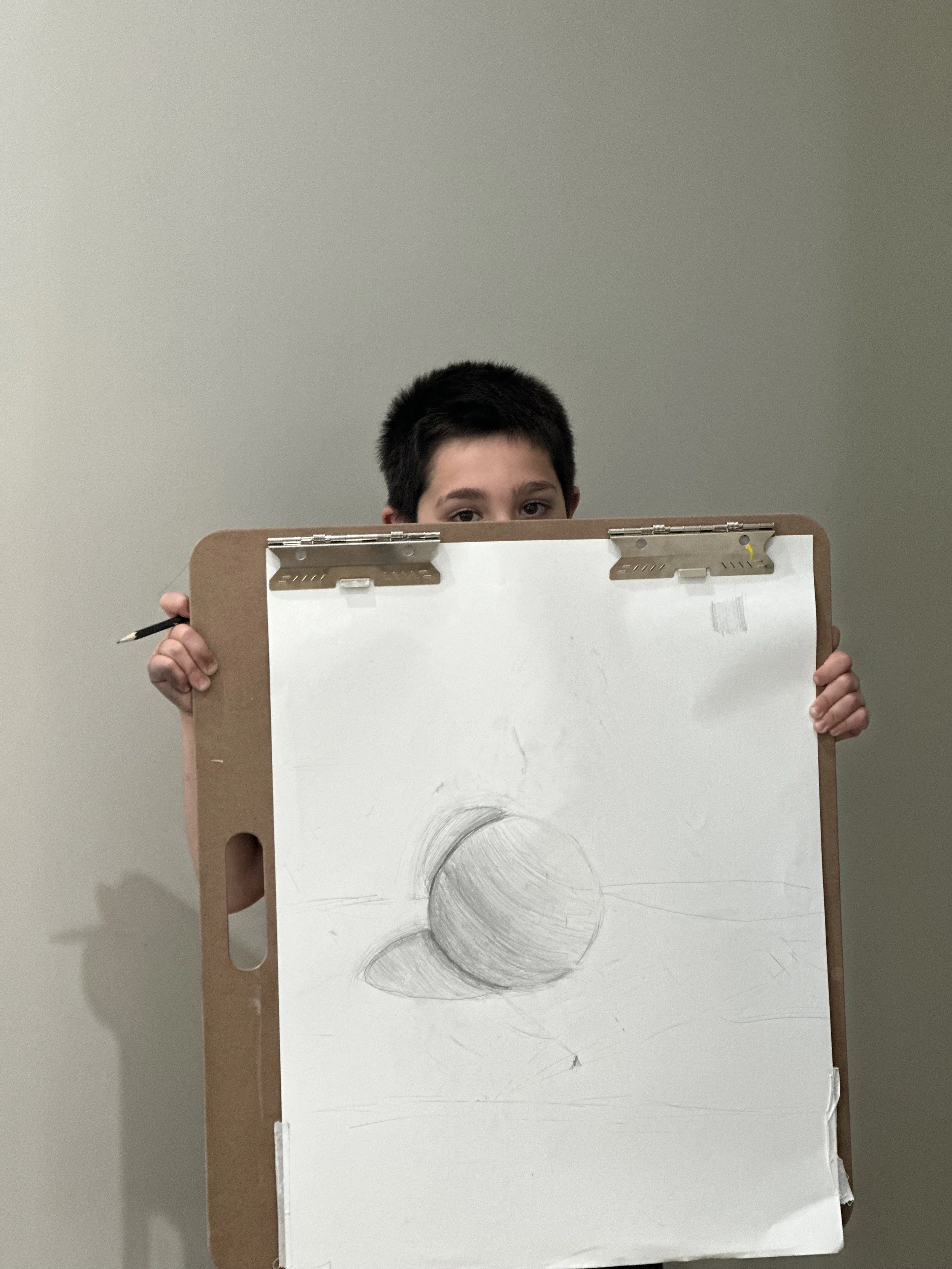 Child with short dark hair peeking over a large sketch pad, holding a pencil, and showing three shaded balls on the paper.