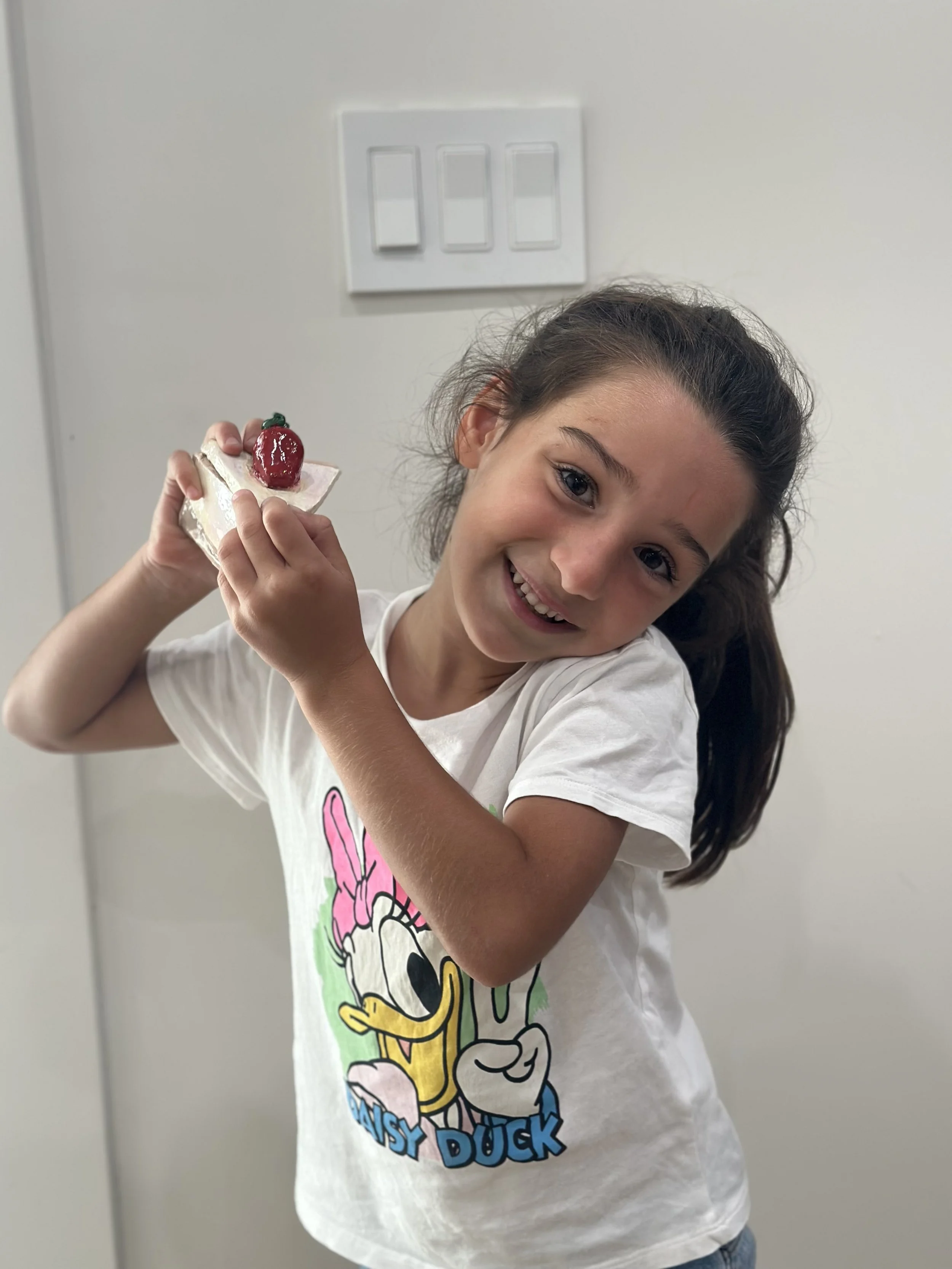 A young girl with brown hair smiling and holding a decorated cake with a strawberry on top, standing in front of a plain white wall with a light switch.