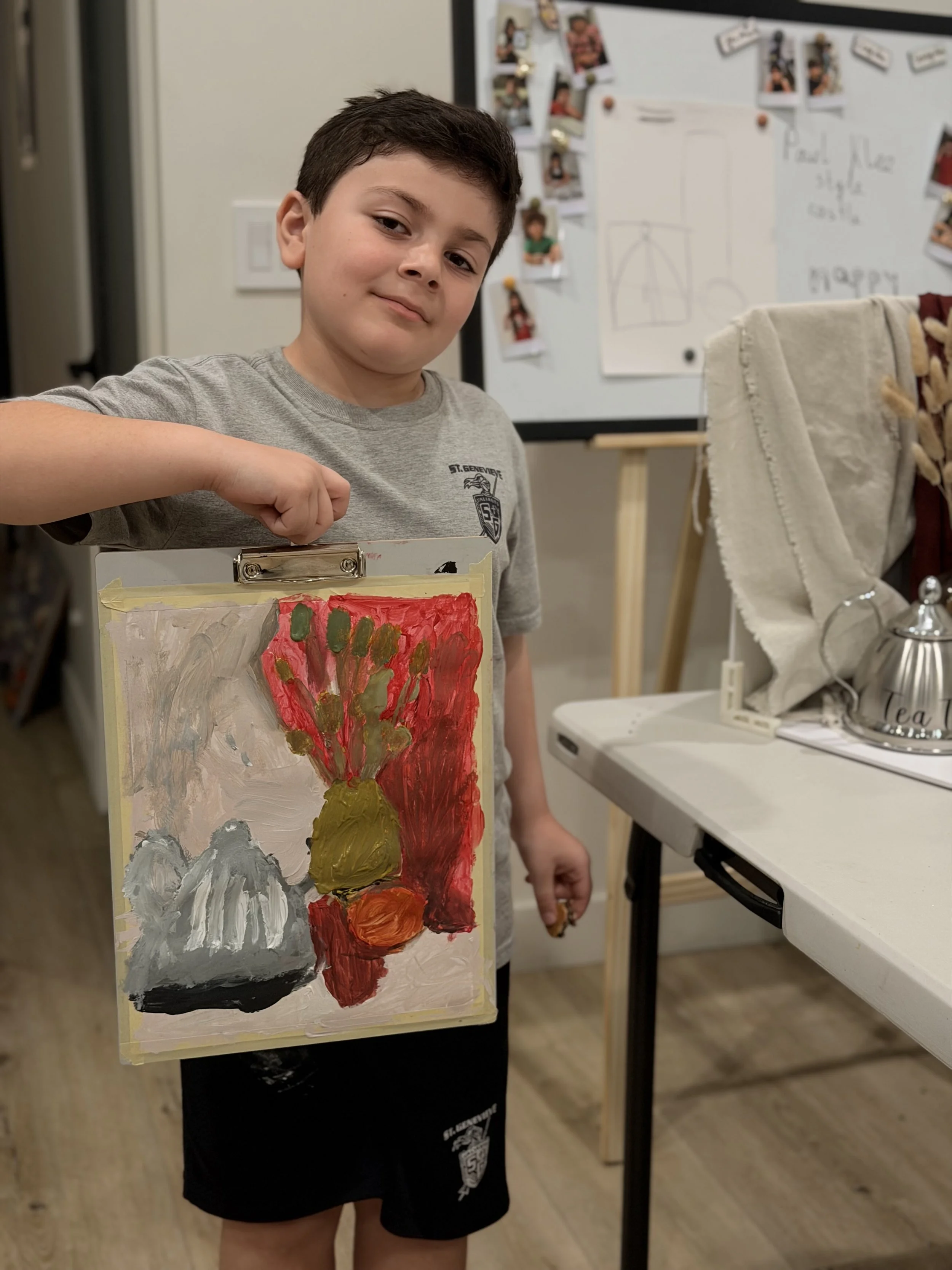 A young boy holding a small canvas painting of a flower with red, yellow, and green colors. The scene appears to be in an indoor art classroom, with a whiteboard and photo collage in the background.