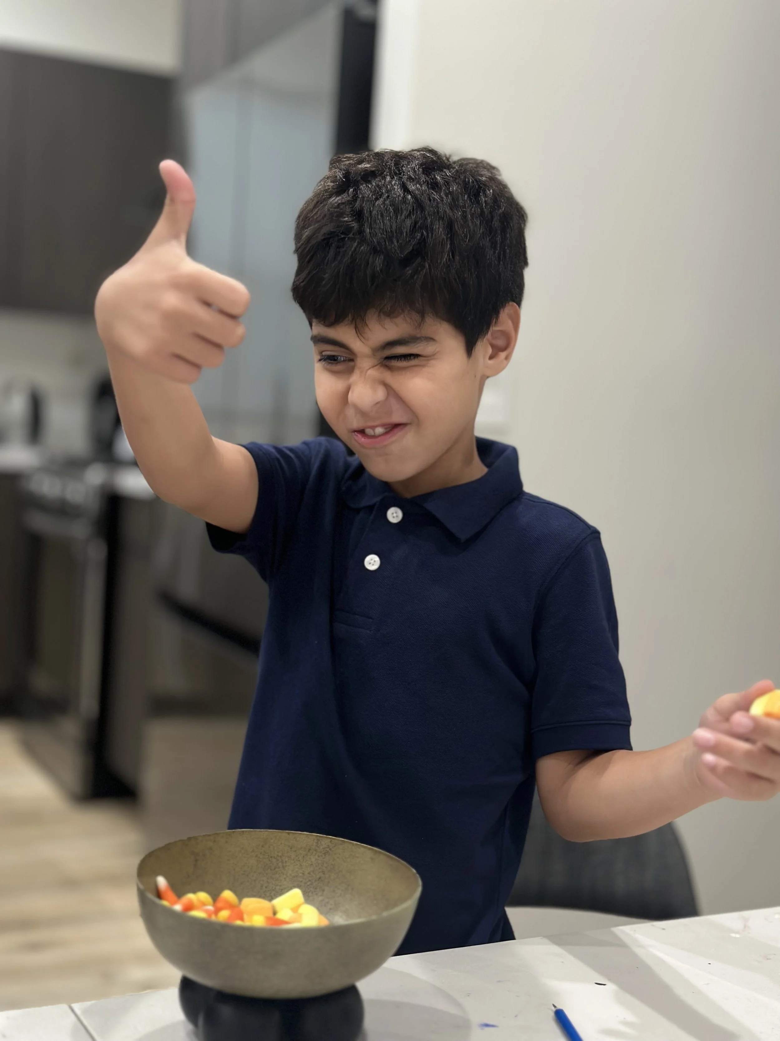 A young boy with dark hair is giving a thumbs up and making a funny face while standing at a table with a bowl of Halloween candy, including candy corn and pumpkin-shaped candies.