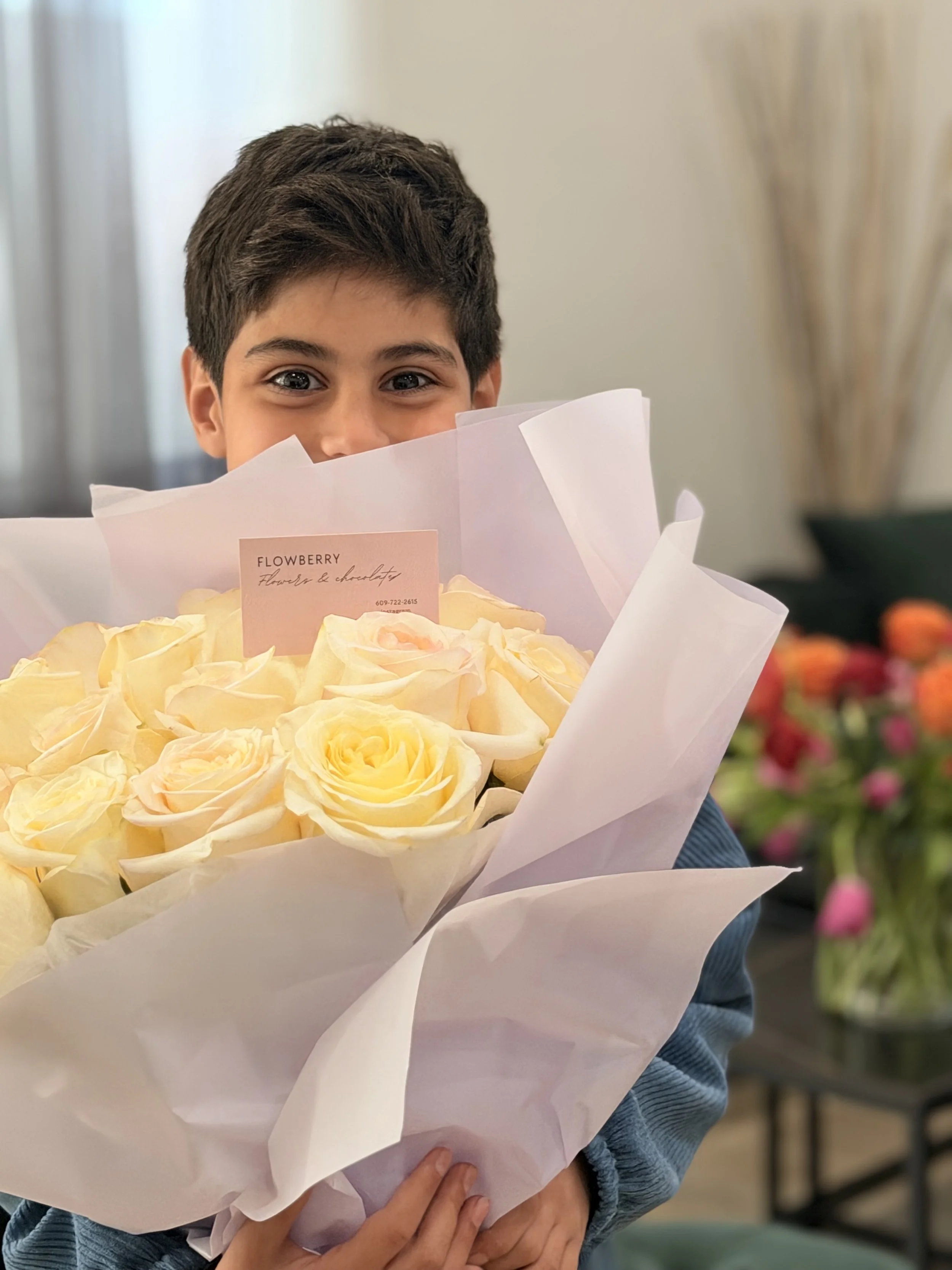 A young boy with dark hair holding a large bouquet of yellow roses wrapped in white paper, with a pink card that reads "FLOWBERRY" in front of him.