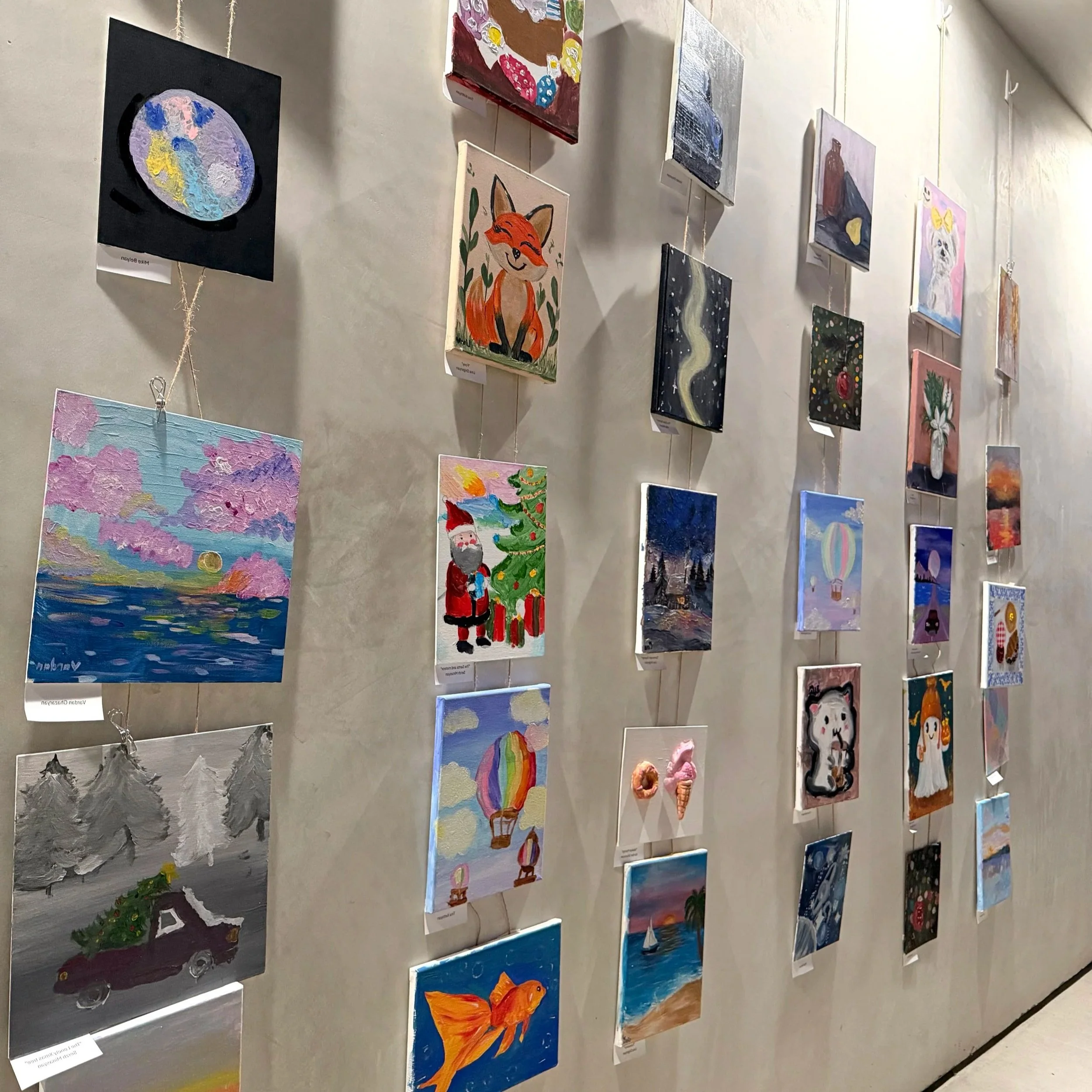 Colorful paintings displayed on a wall in an art gallery, including a fox, a rainbow, a goldfish, and various other scenes.