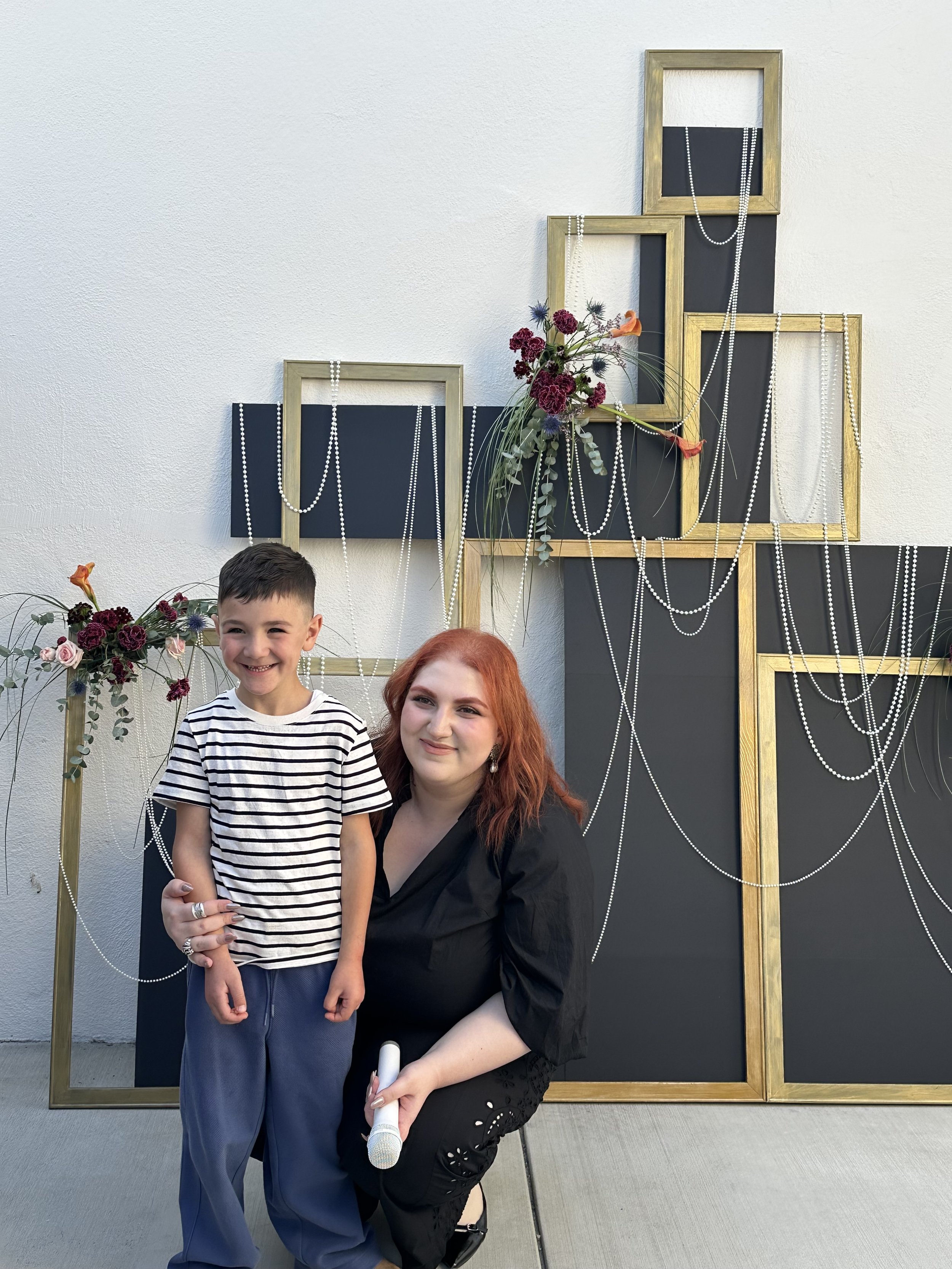 A smiling young boy in a striped shirt standing next to a woman with red hair, both posing in front of a decorative display with black panels, gold frames, white pearl necklaces, and floral arrangements.