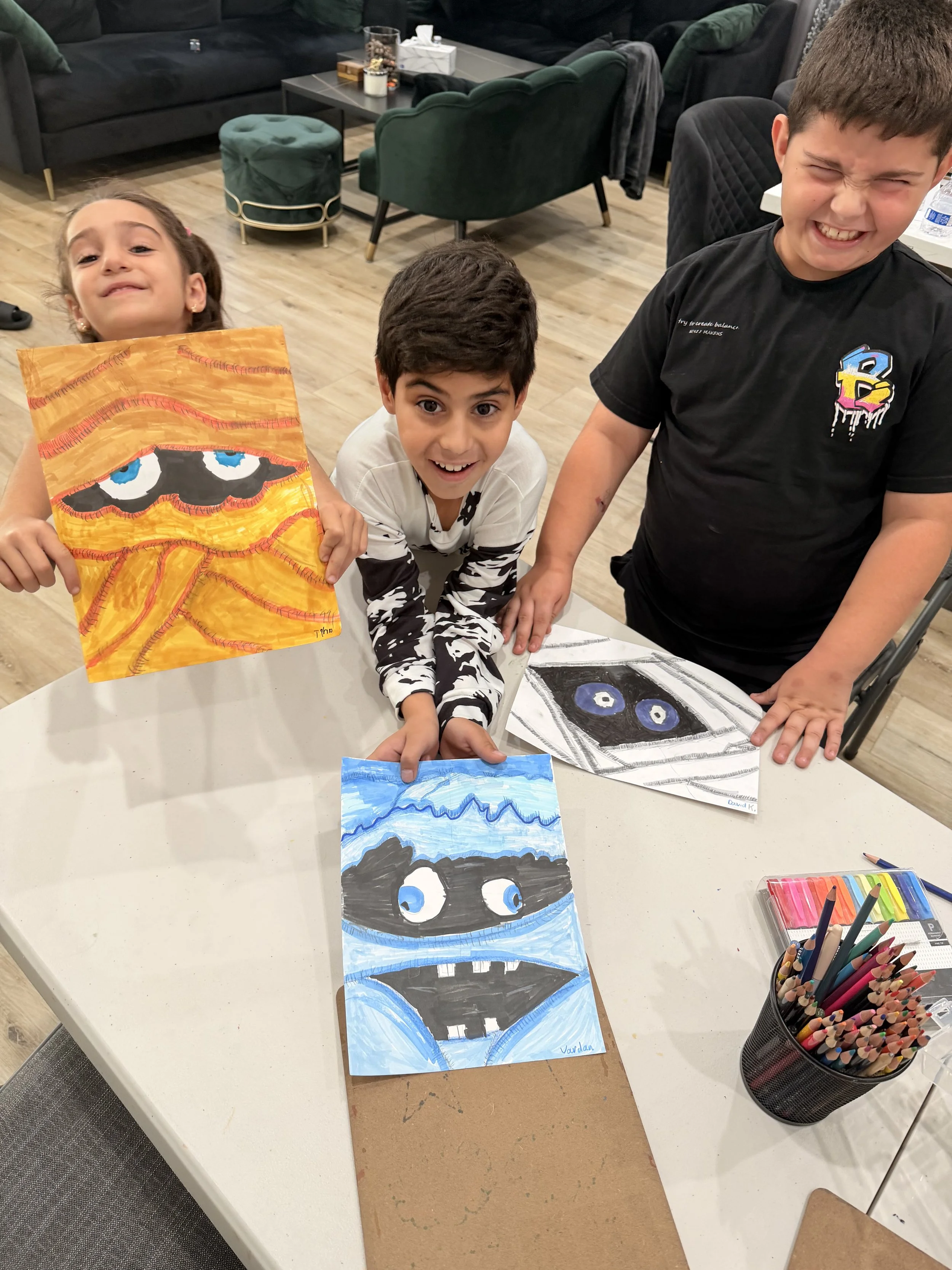 Three children smiling and presenting their colorful paintings of faces with vibrant, bold colors on a table in an indoor space with modern furniture.