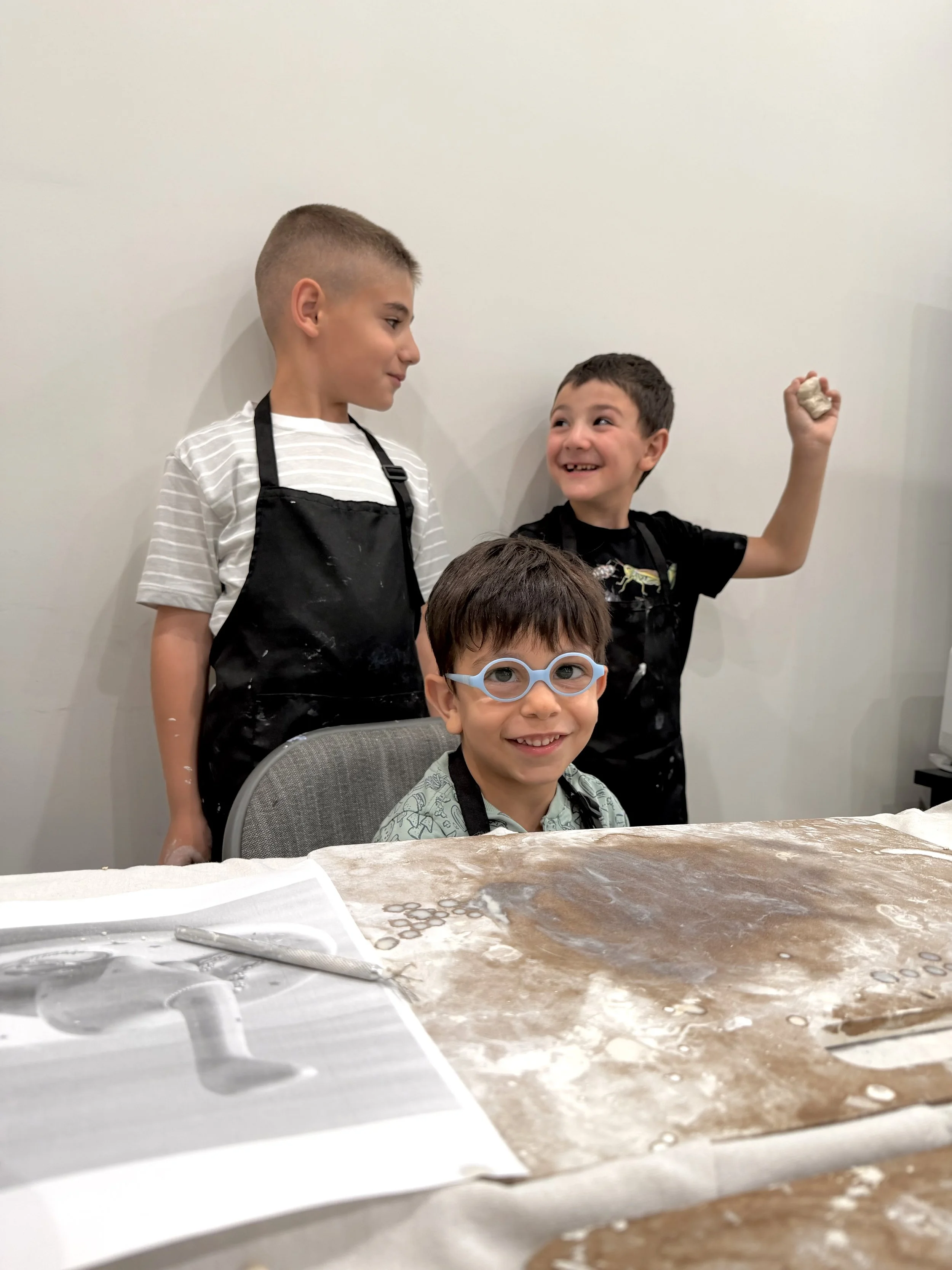 Three young boys at a table with baking supplies in the foreground. One boy is standing, wearing a black apron and striped shirt, looking at another boy who is also in a black apron, smiling and holding a small piece of dough or clay. The third boy, 