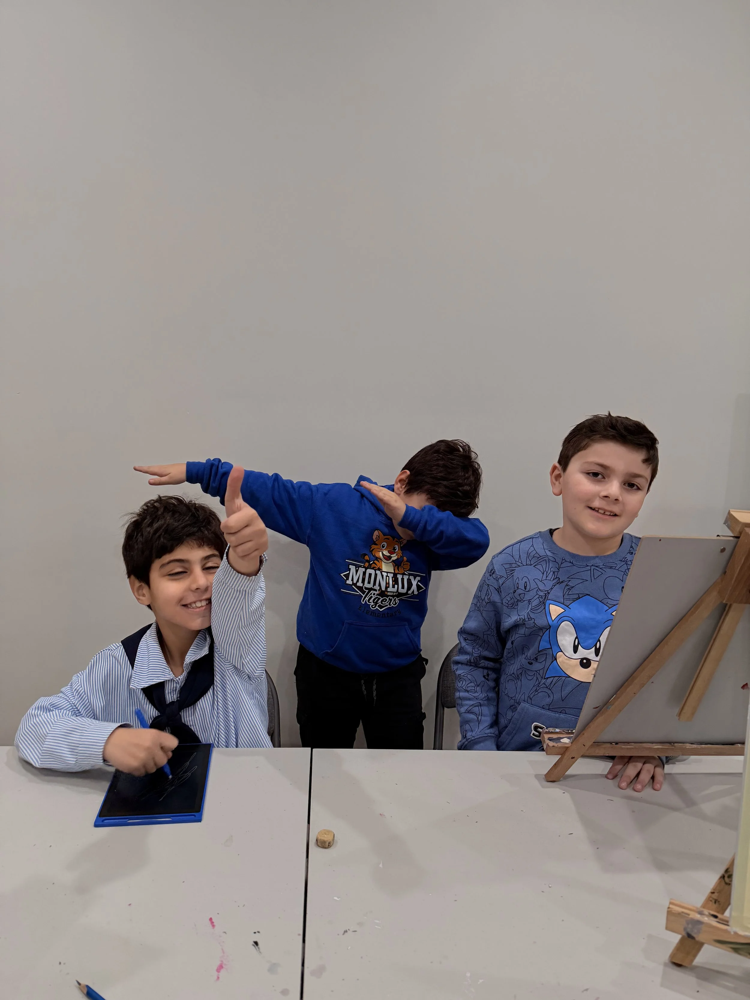 Three young boys are standing behind a white table, with one boy on the left wearing a striped shirt and black backpack, smiling and holding a stylus on a small black and blue drawing tablet. The middle boy is in a blue Monlux Tigers hoodie, dabbing 