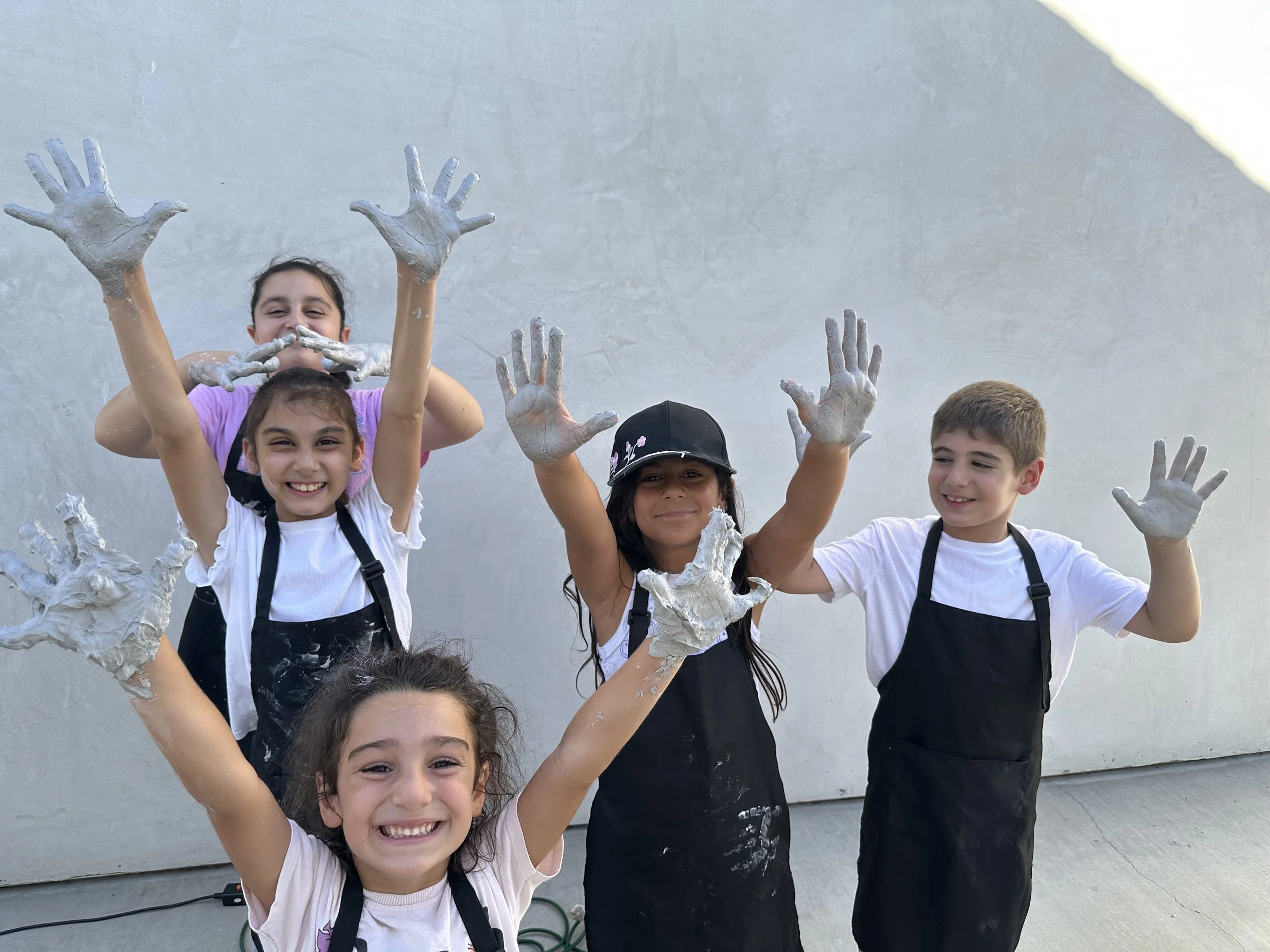 Group of children celebrating with their hands covered in white paint, smiling and showing their painted hands against a plain wall background.