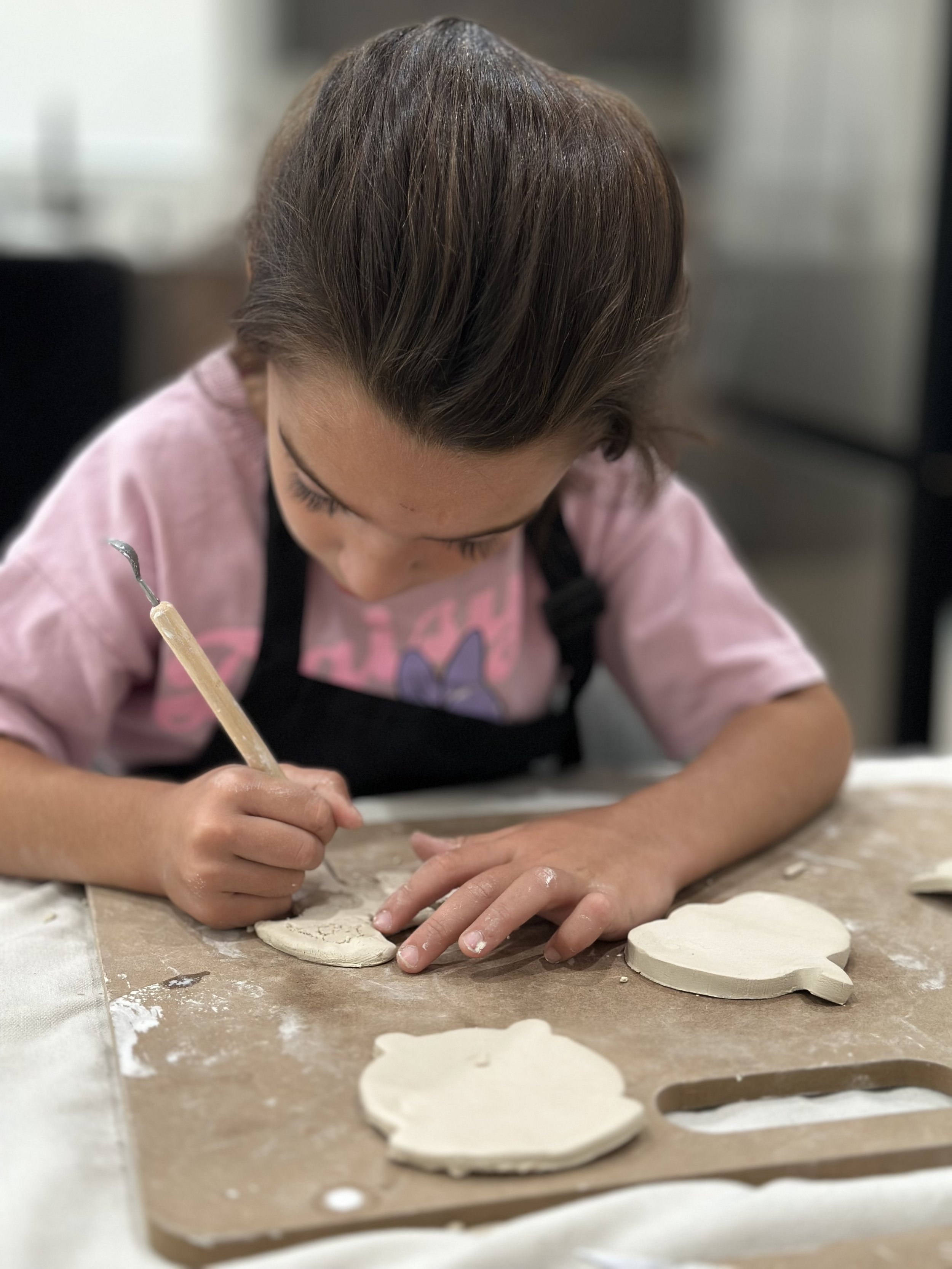 A young girl in a pink shirt and black apron is working on a craft project involving clay or dough. She is using a tool to shape or cut out figures on a flat surface. Several shaped clay or dough pieces are arranged on the table in front of her.