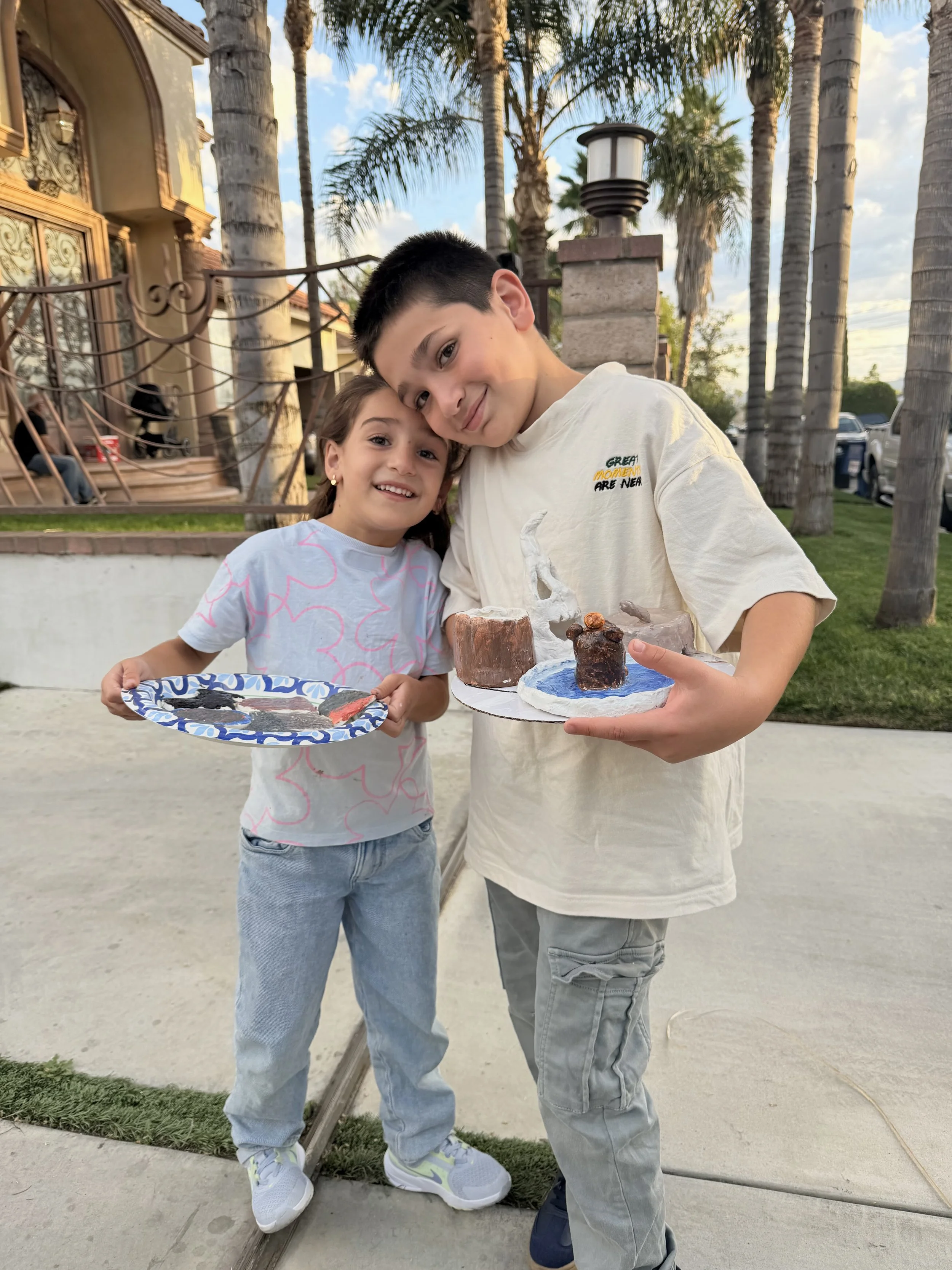 A boy and girl with light skin smiling outside at sunset, holding a craft project with miniature houses. The girl wears a white shirt with pink outlines and blue jeans, and the boy wears a beige t-shirt and gray pants. The background shows palm trees