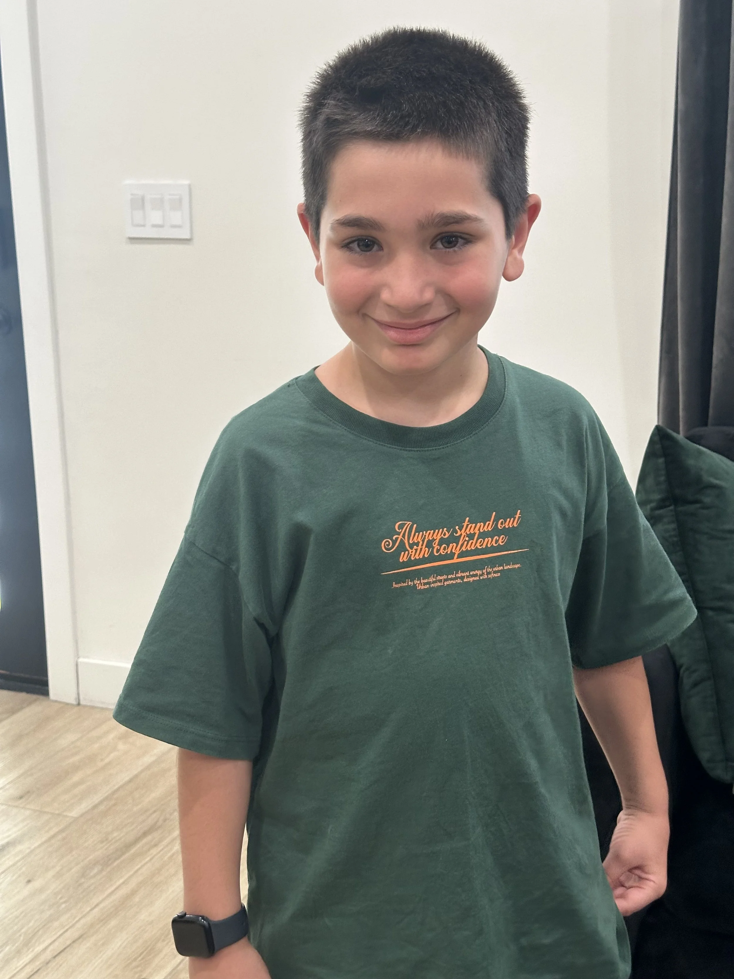 A young boy with short dark hair, wearing a green t-shirt with orange text, smiling at the camera. The background includes a light switch and a black curtain.
