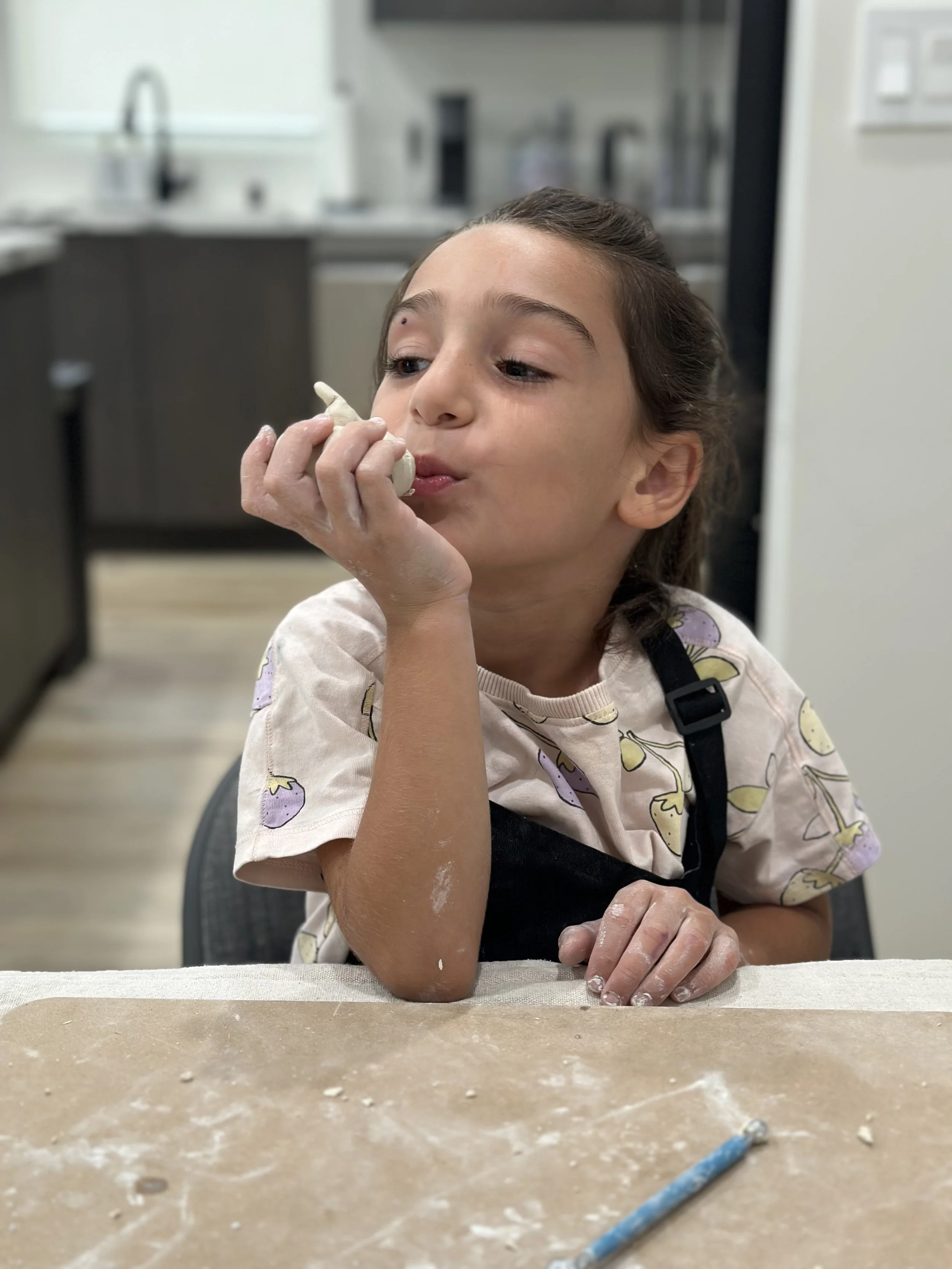 A young girl with long brown hair, wearing a light-colored shirt with a fruit print and a black apron, is sitting at a table in a kitchen. She is holding a small white object near her mouth and appears to be blowing or tasting it, with some flour or 