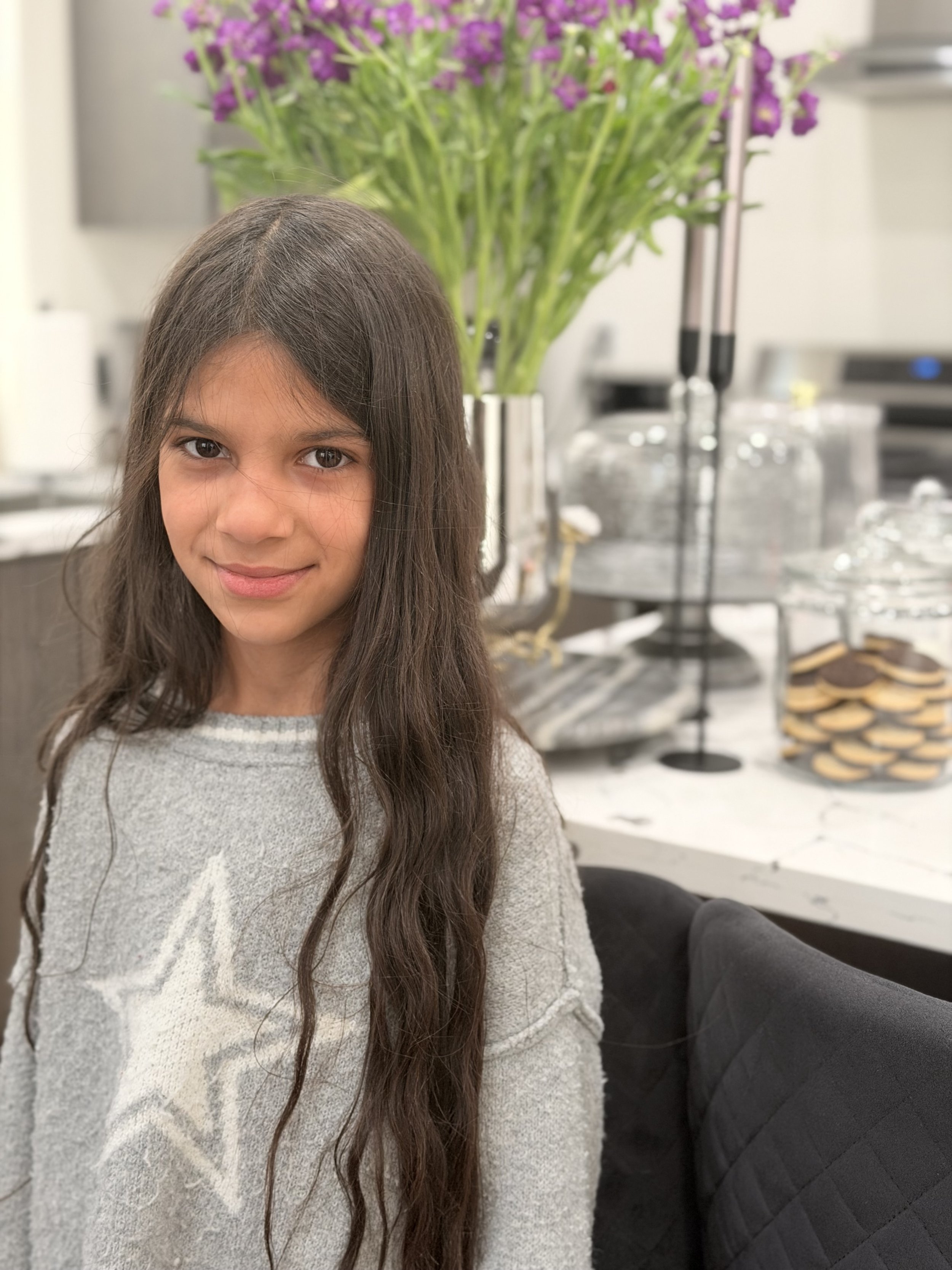 A young girl with long brown hair and a light gray sweater sitting at a table in a room with a large bouquet of purple flowers in a glass vase and a snack tray with cookies and a two-tiered serving stand in the background.