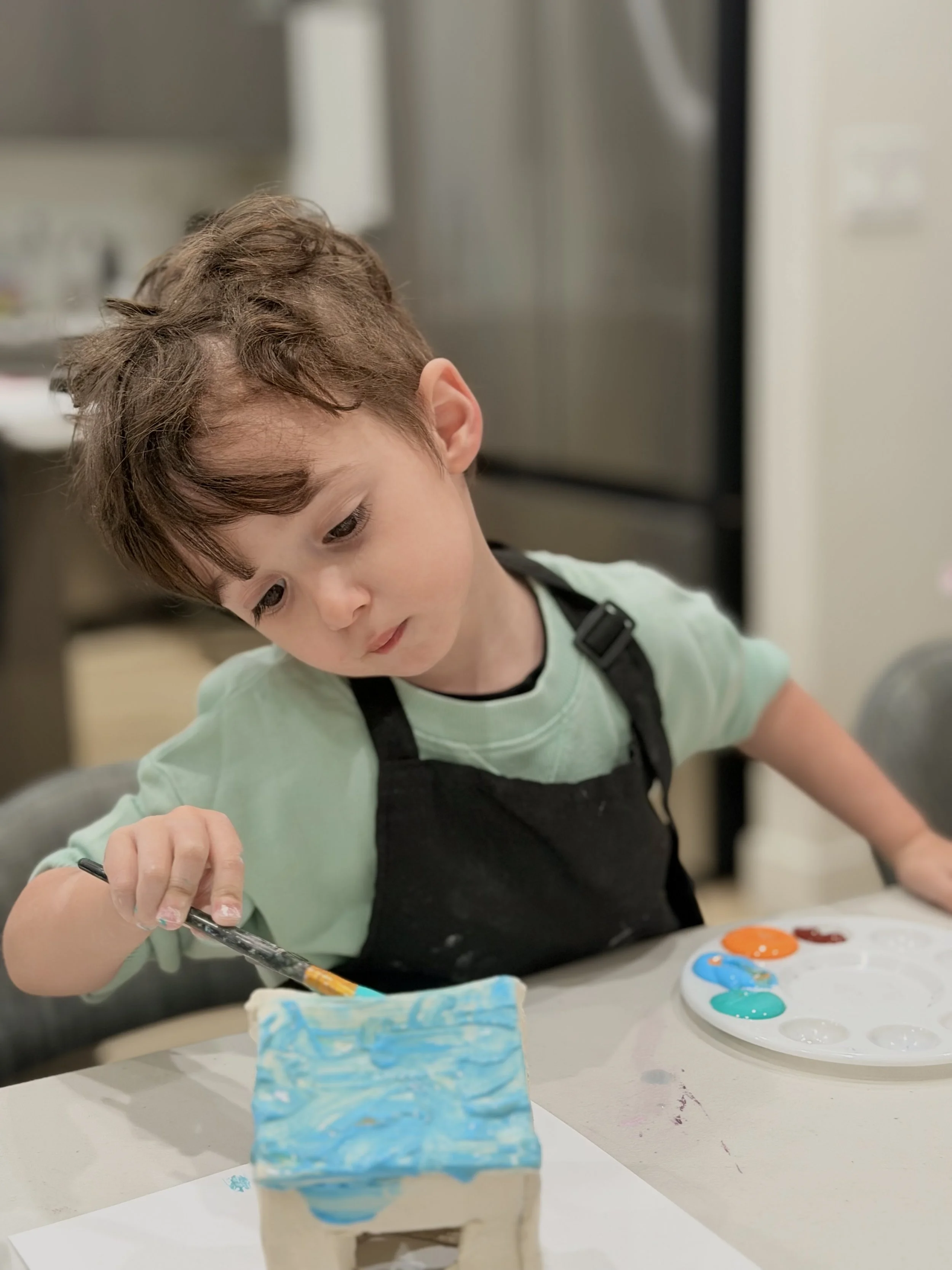 A young boy painting a small ceramic or plaster object with shades of blue, yellow, and orange on a white table, with a palette of paint nearby.