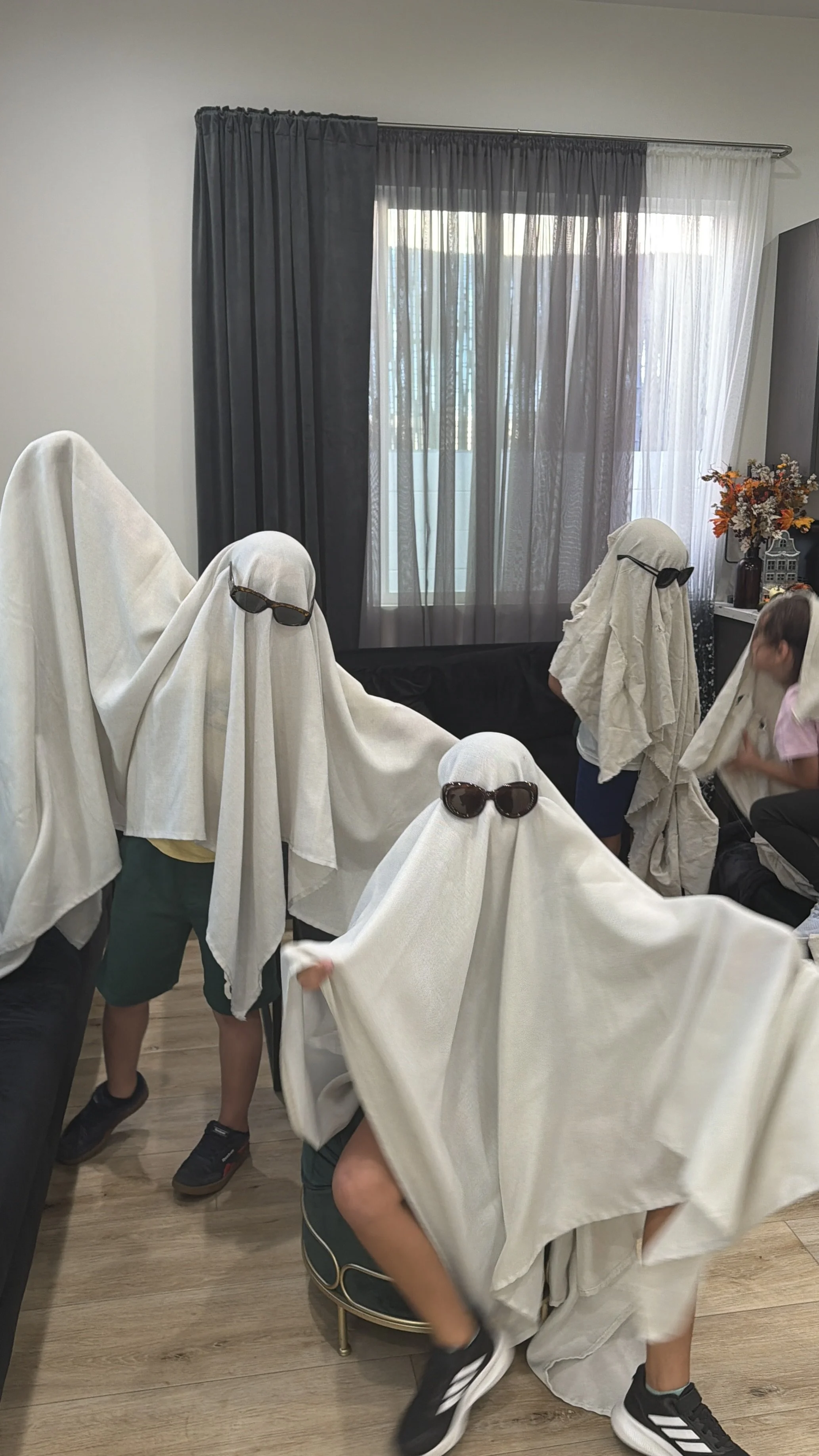 Children wearing white sheets and sunglasses participating in a Halloween or costume event indoors, with some children hiding behind the sheets and others standing or walking around.