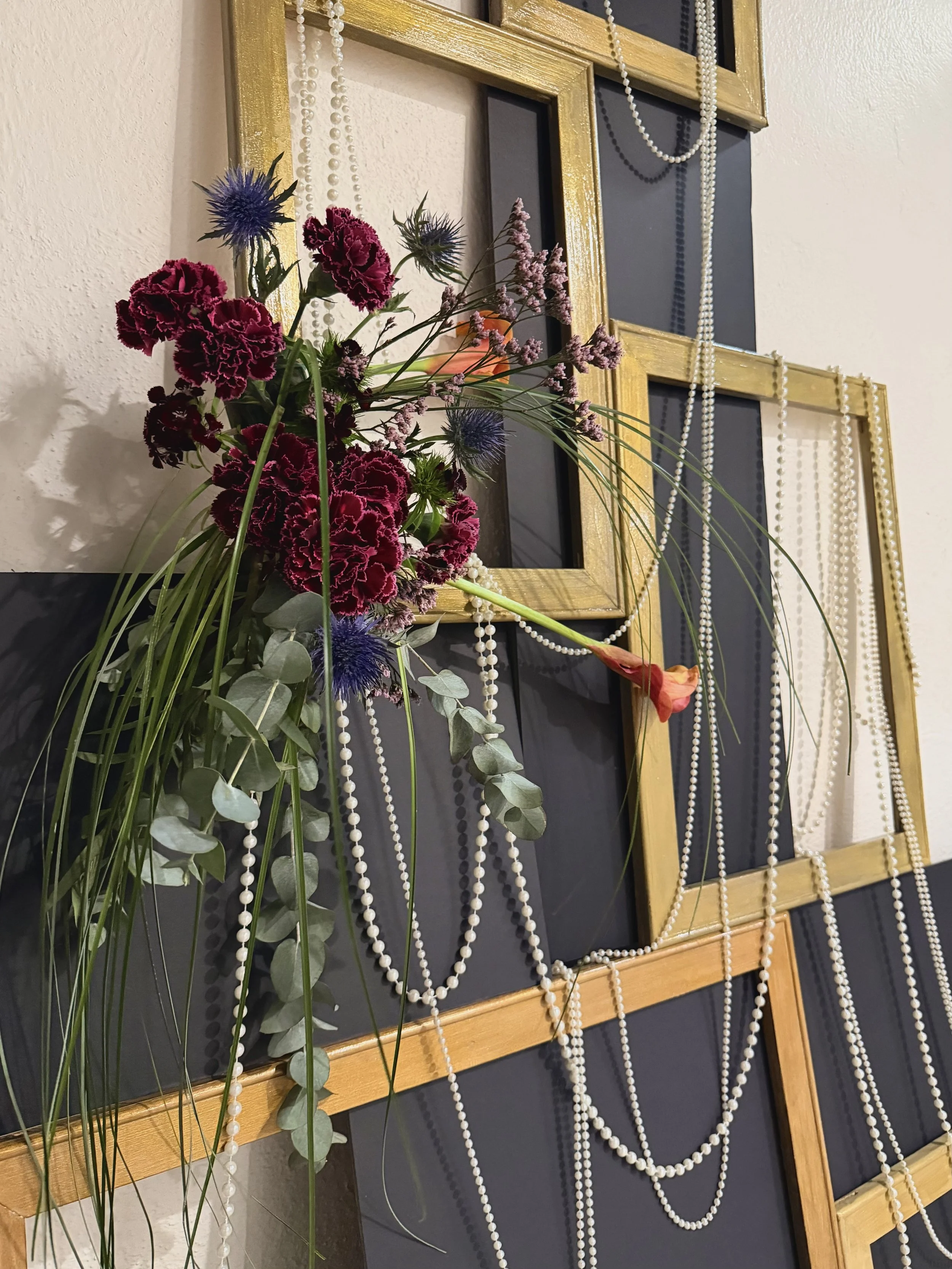 A decorative wall with a gold frame design, black panels, fairy lights, and pearl necklaces, accented with a floral arrangement featuring purple, pink, and green flowers and foliage.
