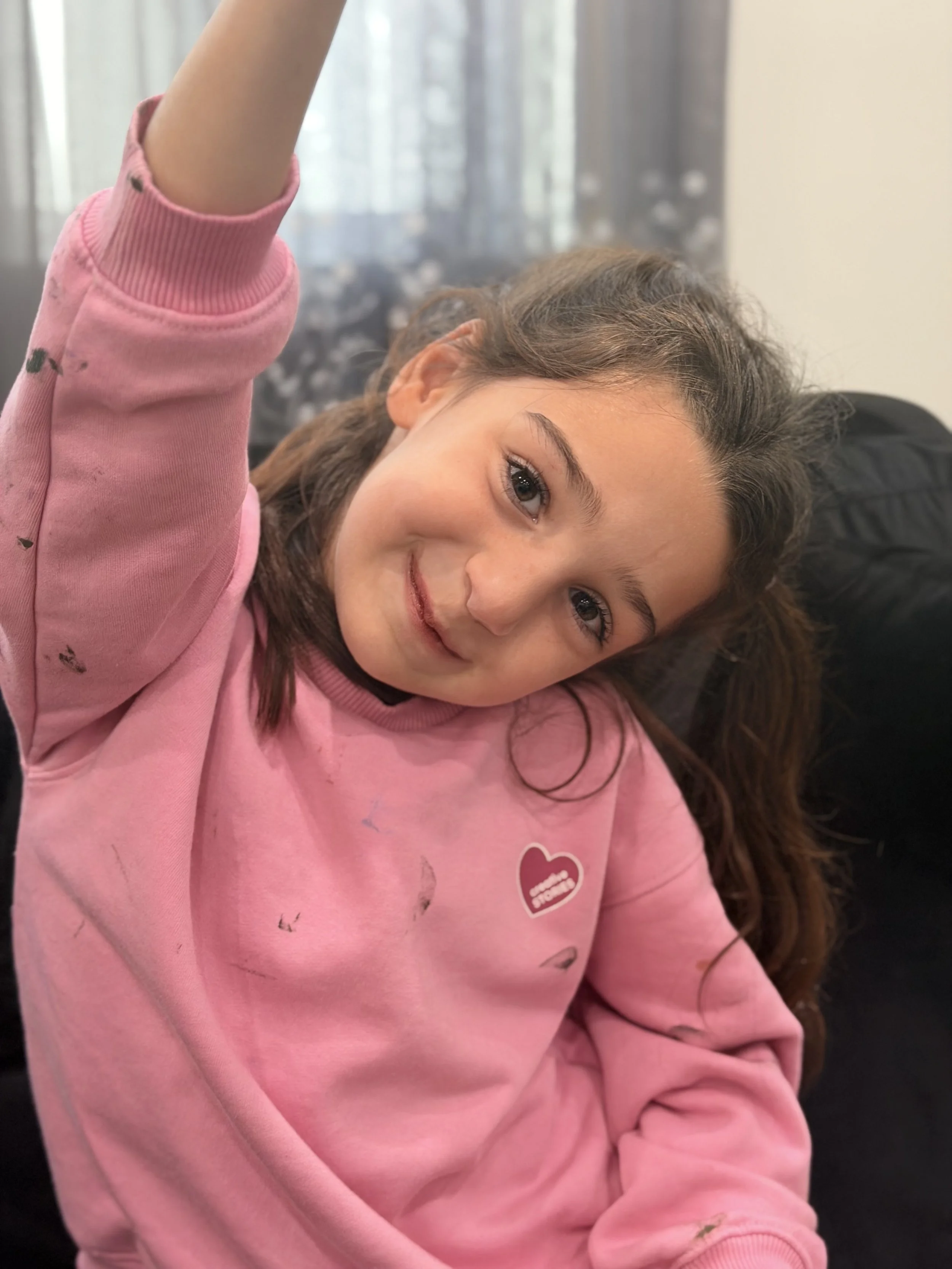 A young girl with long brown hair and brown eyes, wearing a pink sweatshirt, smiling, and resting her head on a black surface.