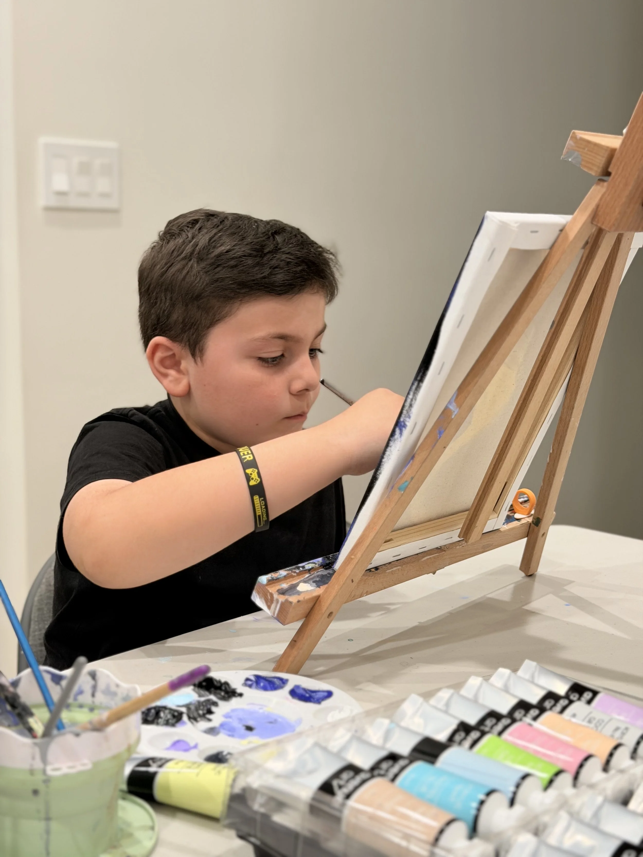 A young boy with brown hair painting on a canvas set on an easel in a room with light gray walls. Various tubes of paint and brushes are on the table in front of him.
