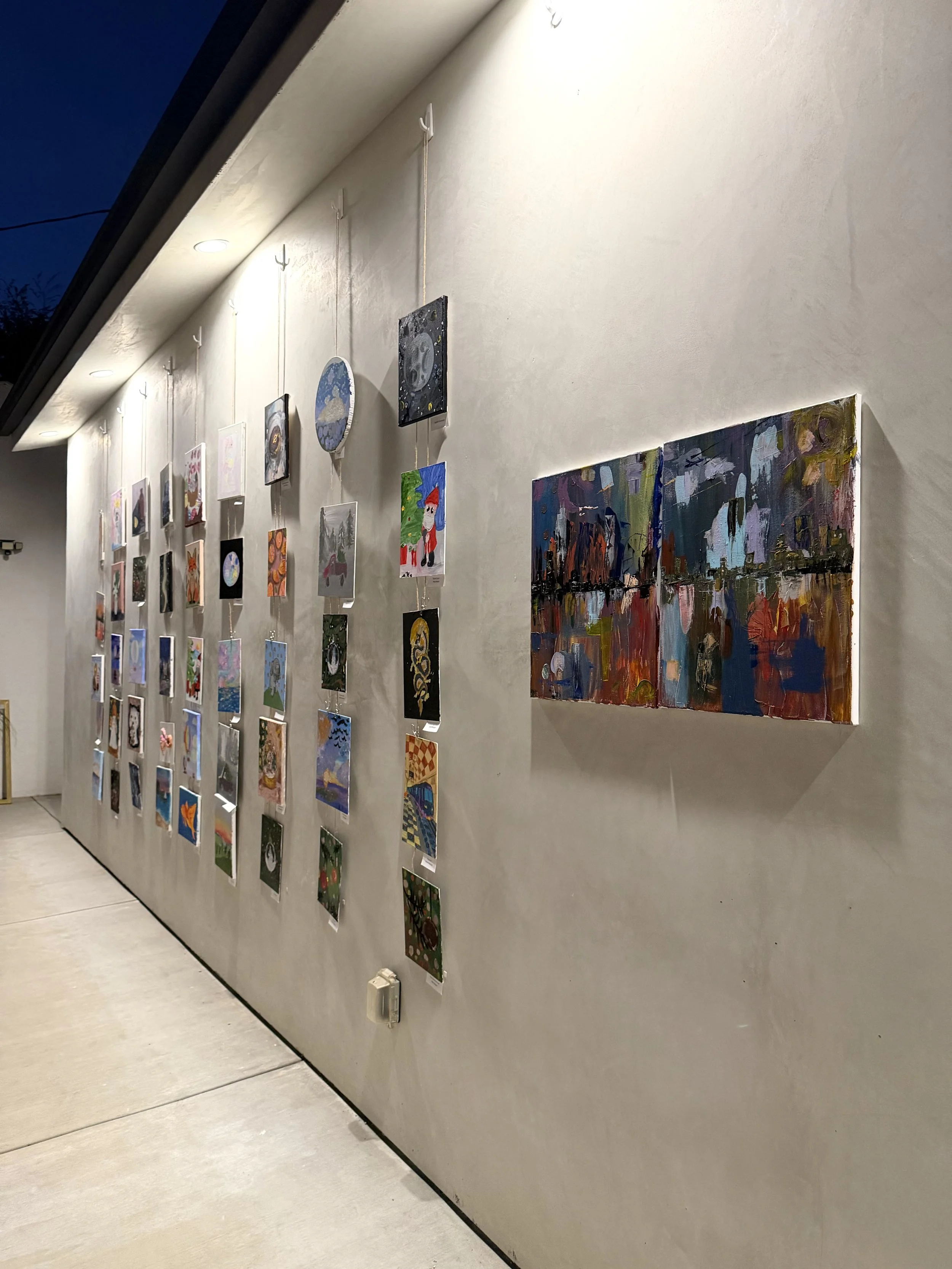 A gallery wall with various small and large colorful paintings and drawings hanging on a white wall, with some items hanging one on top of the other, near the ceiling, in a well-lit indoor space.