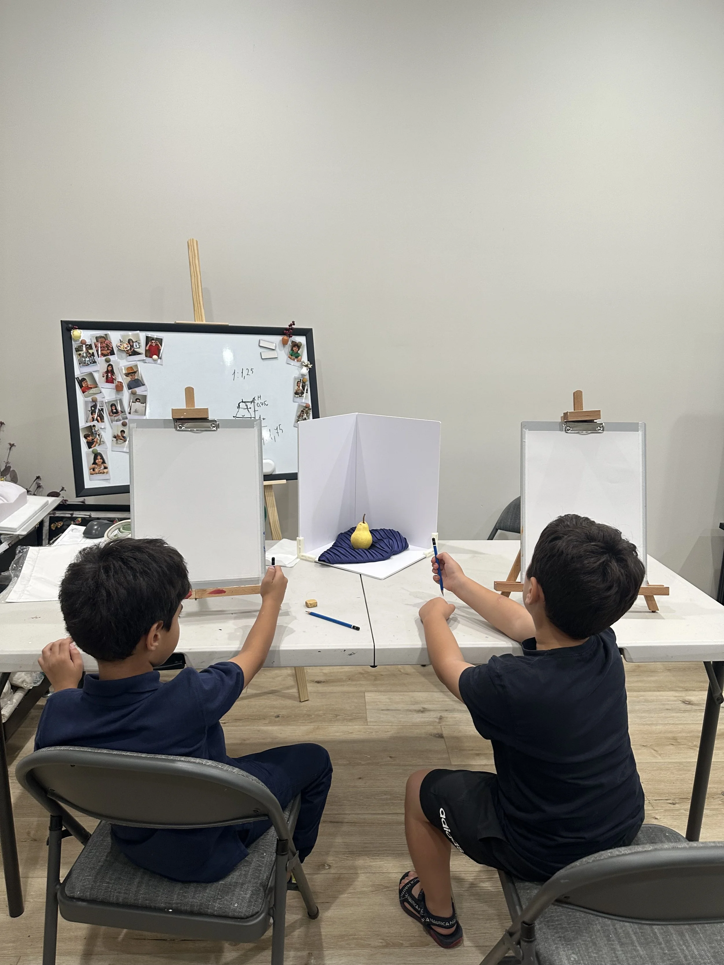 Two children sit at a table, working on a project with whiteboards, markers, and a small pear prop, in a room with a plain wall and wooden floor.