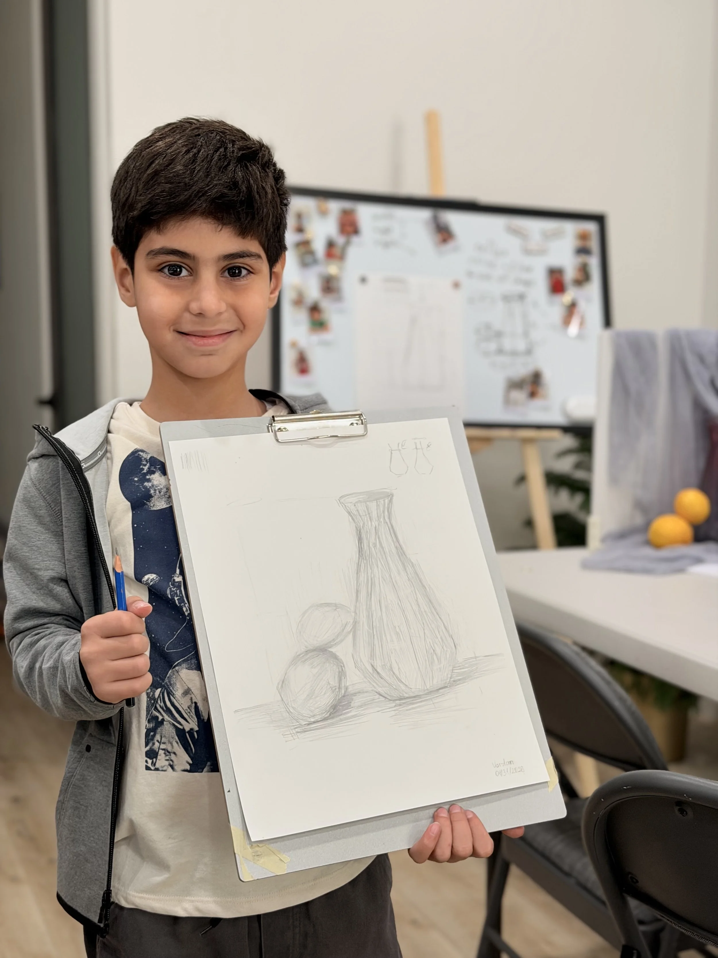 A young boy with dark hair and a smile holding a drawing of a vase with round objects on top. He is standing indoors in front of a blurred background with a whiteboard and some yellow lemons on a table.