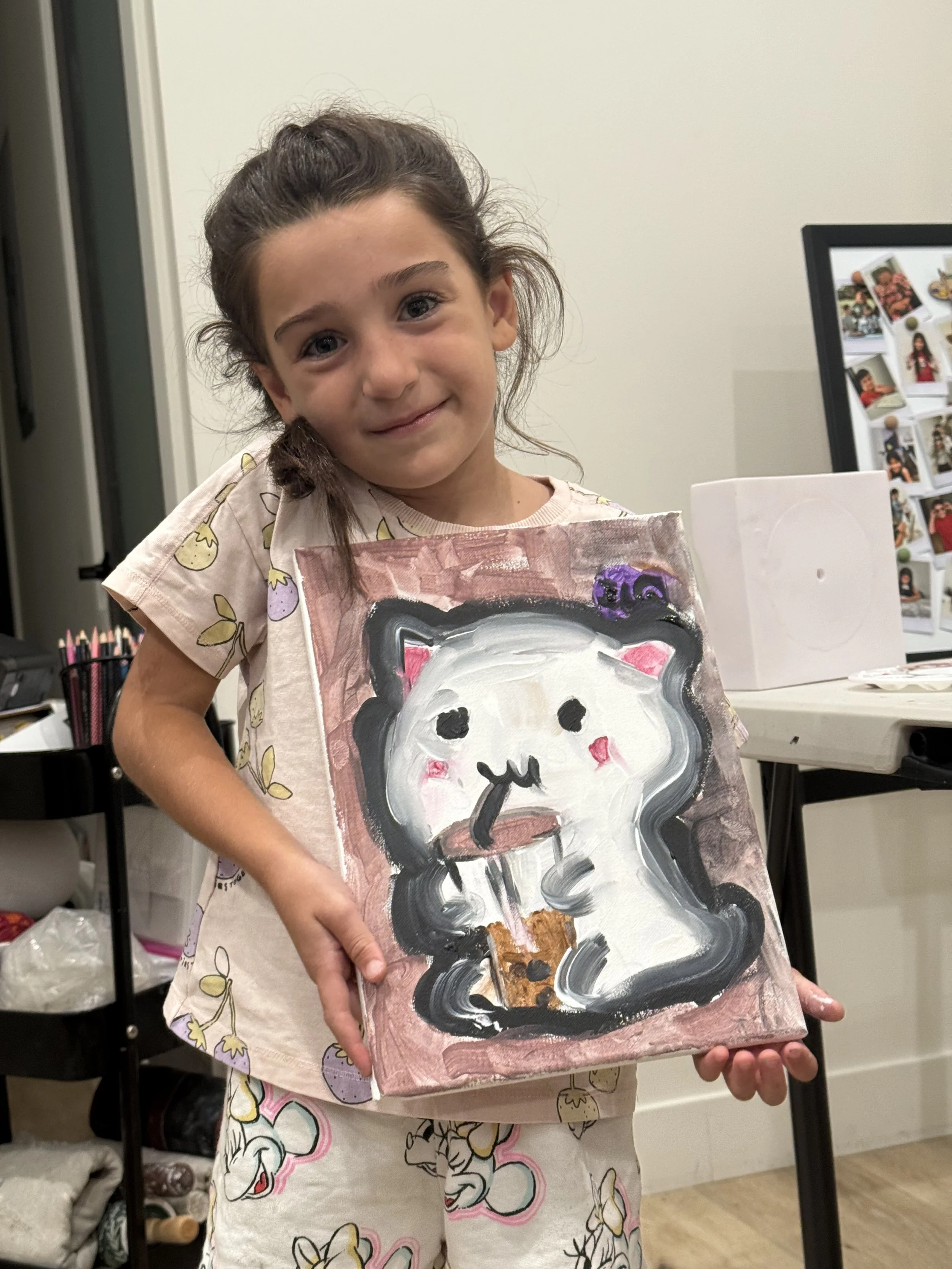 A young girl with brown hair and dressed in pajamas with a floral pattern, standing indoors, holding a painted canvas of a cartoon snowman with blush cheeks, a black hat with purple swirls, and a brown scarf. She is smiling slightly and looking at th