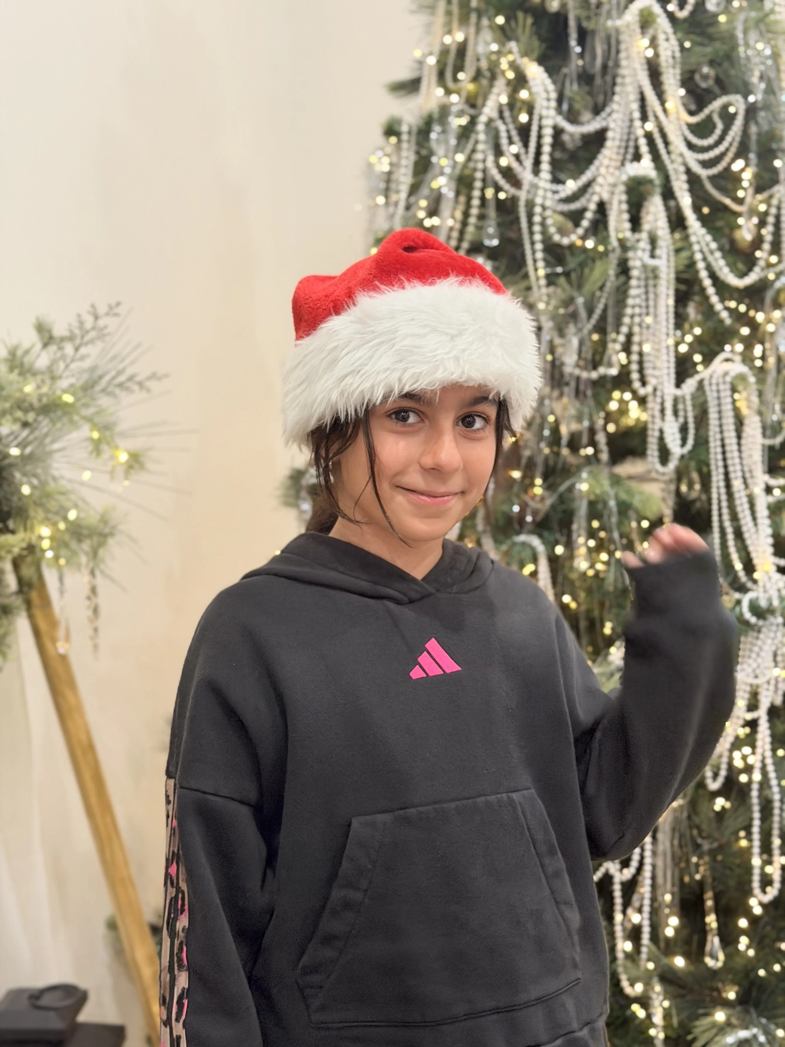 A young girl wearing a red and white Santa hat and a black hoodie with pink accents, standing in front of a decorated Christmas tree with white beads, lights, and ornaments.