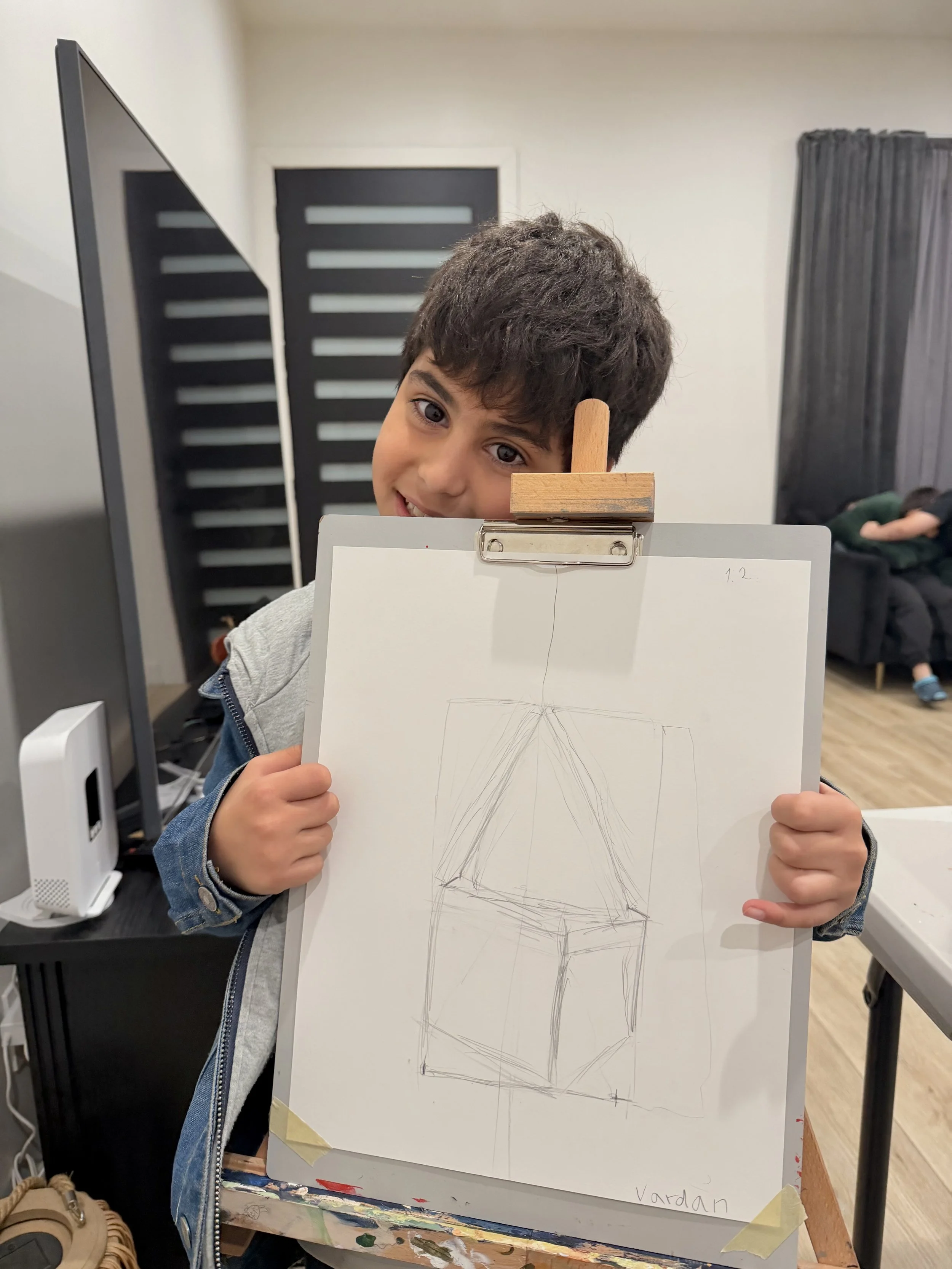 A young boy with dark curly hair smiling and holding up a whiteboard with a rough pencil sketch of a 3D cube drawn on it. The boy is standing indoors near a black computer monitor and a black privacy screen, with a person sitting on a couch in the ba