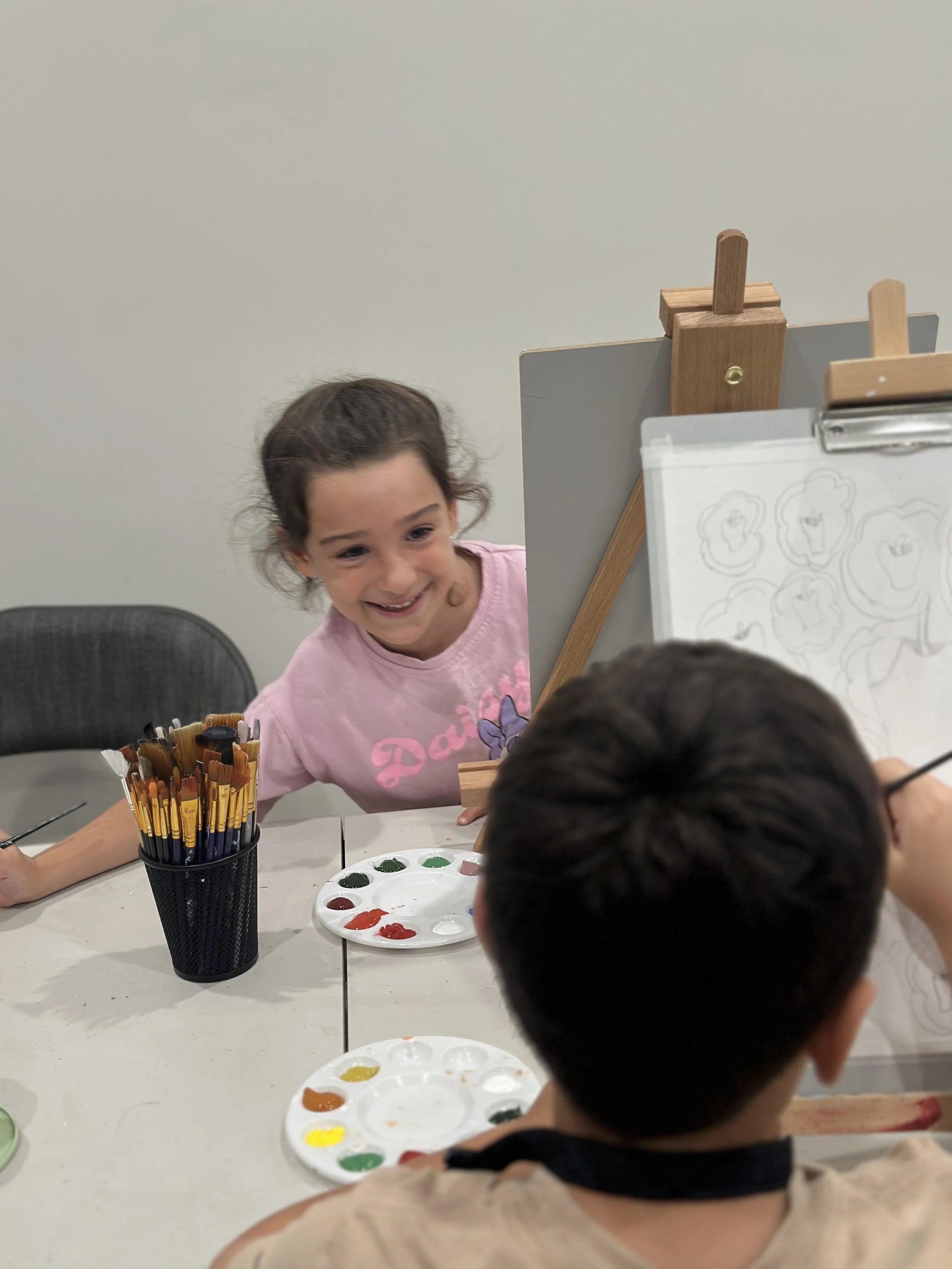 Young girl smiling at a painting activity with paint palette, brushes, and watercolors on a table.