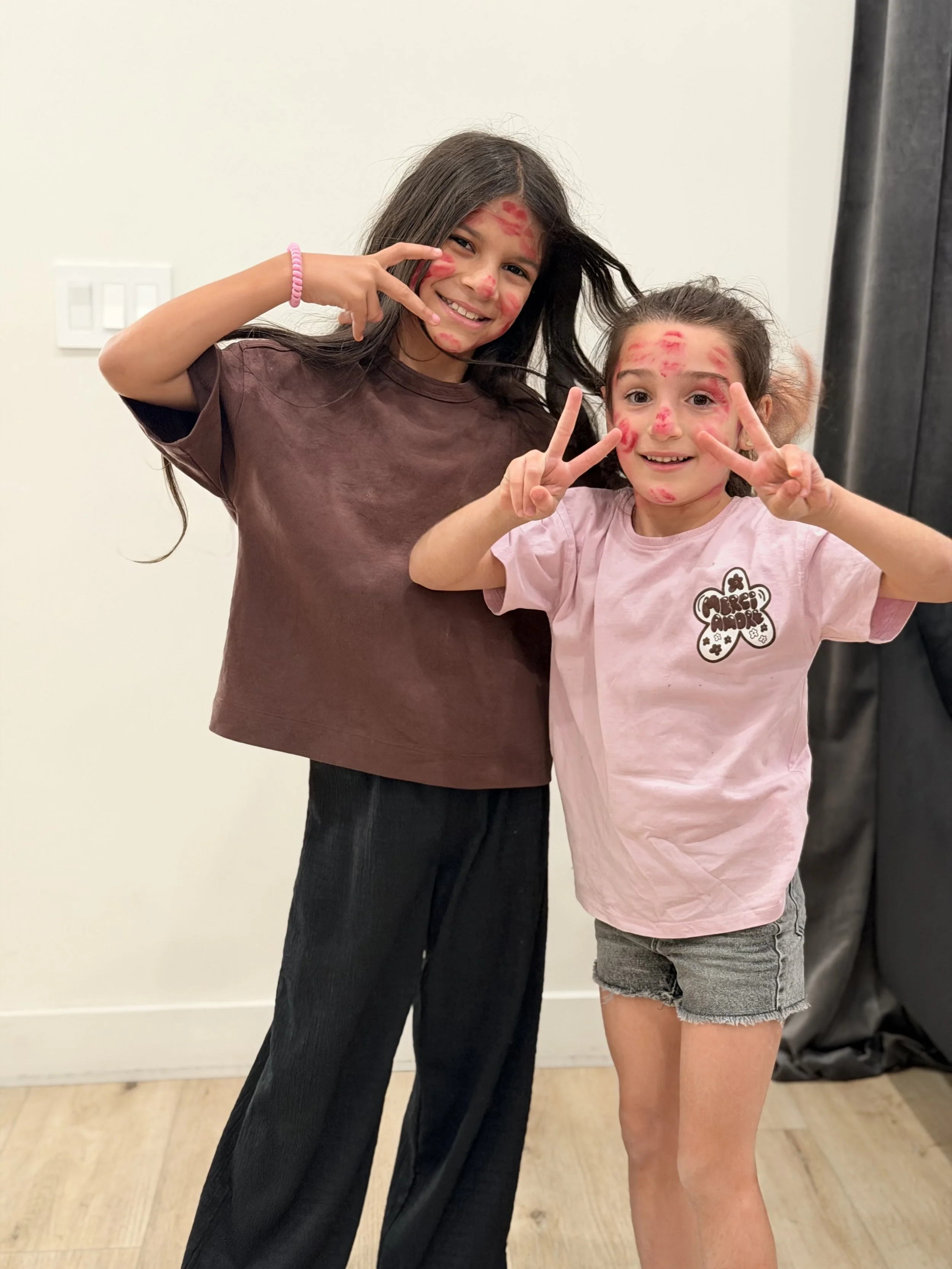 Two young girls with painted faces making peace signs and smiling indoors against a plain white wall.