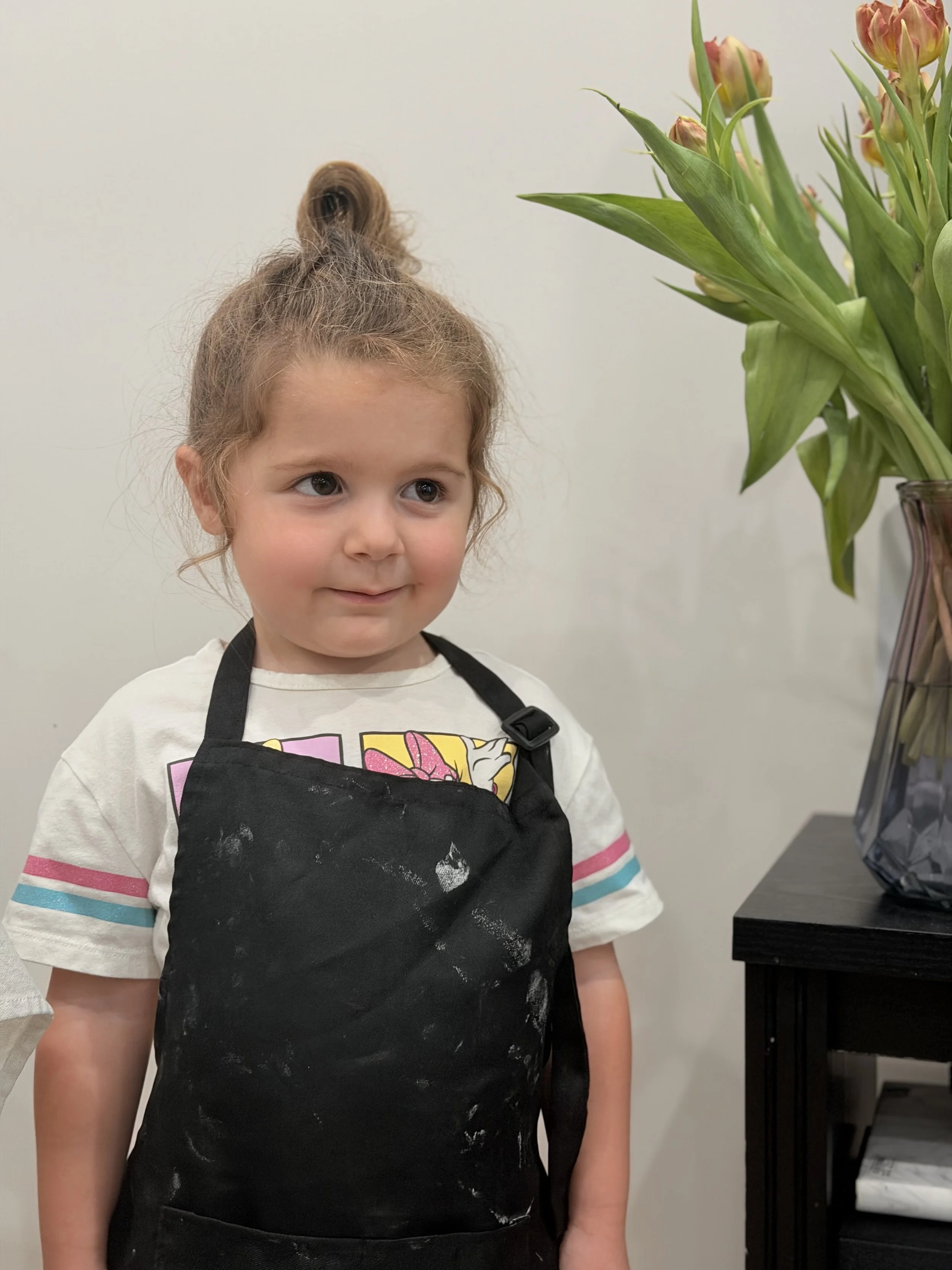 A young girl with light skin, brown hair tied in a high bun, wearing a white t-shirt with colorful stripes near the sleeves, and a black apron with white paint stains, standing beside a vase of flowers, looking to her left with a slight smile.